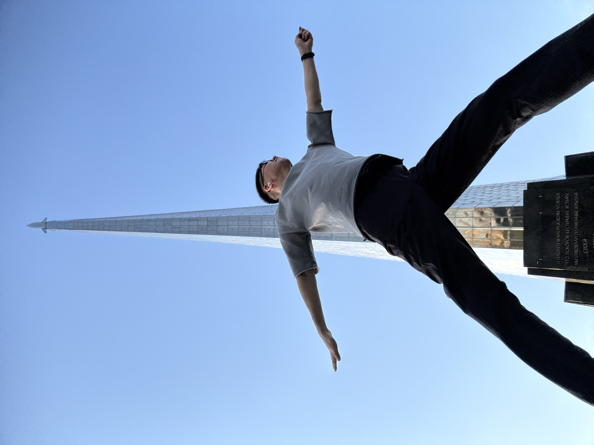 A low-angle medium shot captures a young man in a grey t-shirt and black pants with arms outstretched against a clear blue sky. The man is positioned in the foreground, with a towering silver monument resembling a rocket ship behind him, dominating the background. The monument appears to be the Monument to the Conquerors of Space, located in Moscow, Russia. The man's body is angled, his left arm extended to the side, and his right arm bent at the elbow with his fist clenched. His head is tilted back, and he appears to be looking upwards towards the monument or the sky. The base of the monument is visible, with inscriptions on a dark granite slab. The scene is illuminated by daylight, casting soft shadows.