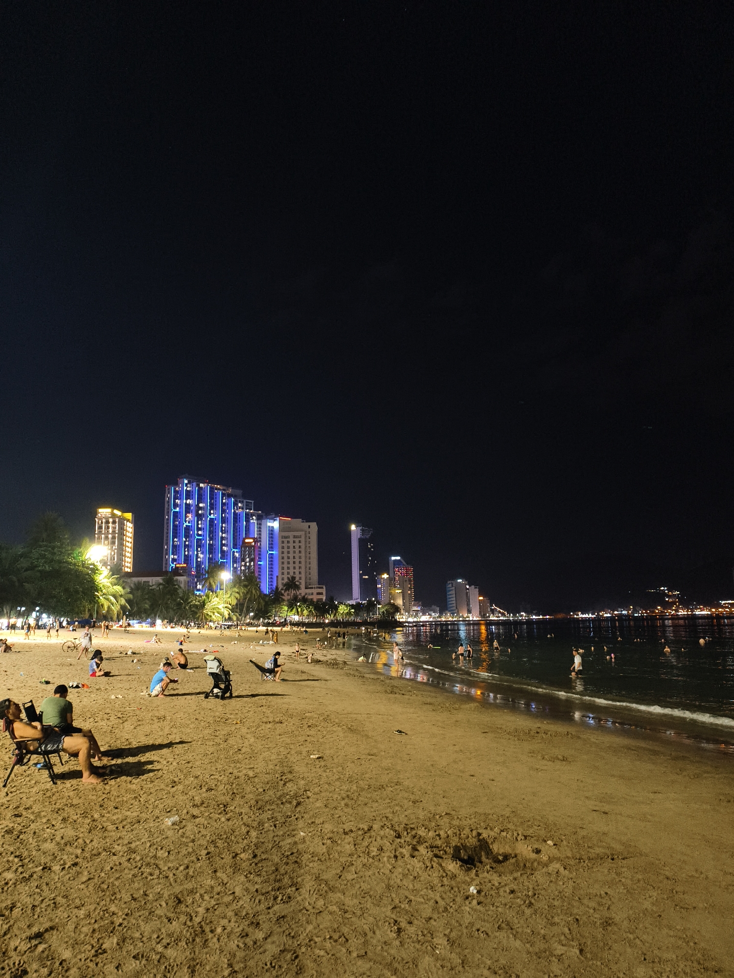 At night, a sandy beach in Nha Trang city, Vietnam, is populated by numerous individuals and groups. Several people are seated on chairs on the sand, while others are walking or lounging. In the water, some individuals are swimming or wading. The shoreline is illuminated by the lights of a city skyline featuring tall buildings with colorful external lighting, including a prominent structure with blue lighting. Palm trees are visible along the edge of the beach, and the ocean reflects the city lights. The sand itself shows evidence of human activity with scattered debris.