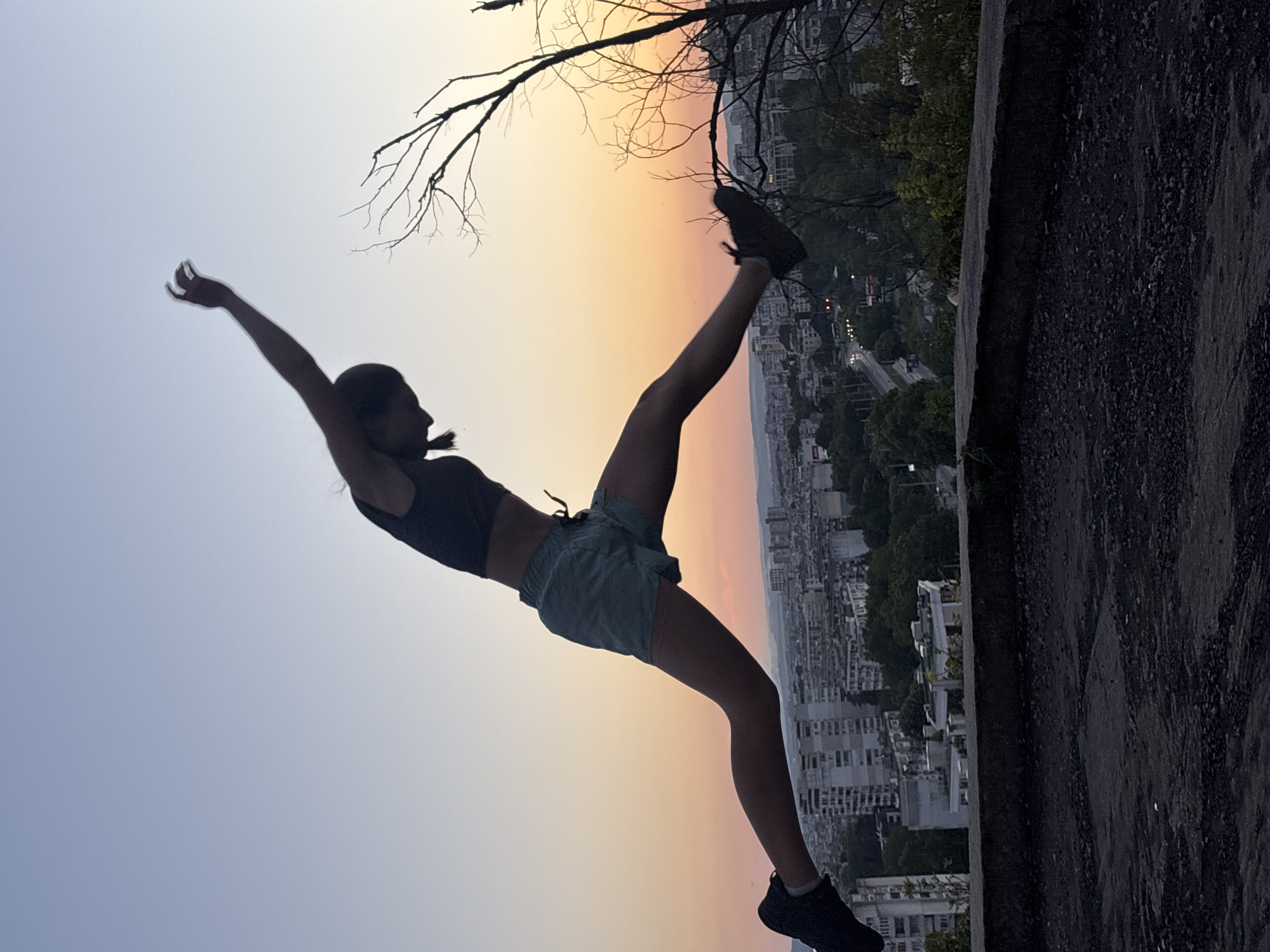 A silhouette of a young person is captured mid-air in a split jump against a gradient sky of soft oranges and blues, indicating sunset. The person is wearing a dark sports bra and light-colored shorts, with dark athletic shoes. Their arms are outstretched, one upwards and the other downwards, contributing to the dynamic pose. The background reveals a city skyline, identifiable as Varna, Bulgaria, with buildings and trees visible on a hillside. A dark, textured surface, possibly a concrete ledge or rooftop, is in the foreground. A bare tree branch extends from the right side of the frame, adding to the natural elements of the scene. The overall impression is one of freedom and athleticism in an urban setting during twilight.