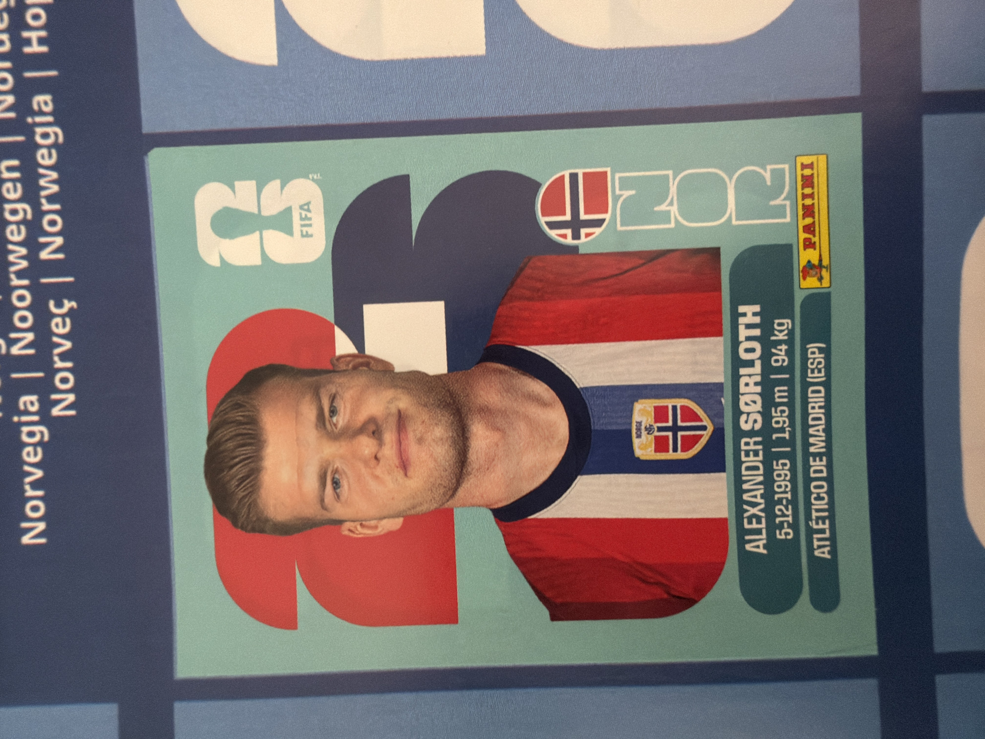 The image shows a close-up of a football sticker featuring Alexander Sørloth. The sticker has a teal background with large red and blue numbers "23" behind the player. Sørloth is wearing a red and white striped jersey with a dark blue collar, and a crest on the chest with the word "NORWAY" above a Norwegian flag. Below his image, his name "ALEXANDER SØRLOTH", his birthdate "5-12-1995", height "1,95 m", weight "94 kg", and club "ATLÉTICO DE MADRID (ESP)" are printed. A yellow "PANINI" logo is present in the bottom right corner of the sticker. The sticker is part of a larger collection, with a portion of the word "Norvegia" visible at the top of the image, and other stickers or graphics to the right and left. The overall image is a photograph taken from a slightly elevated angle, focusing on the sticker.