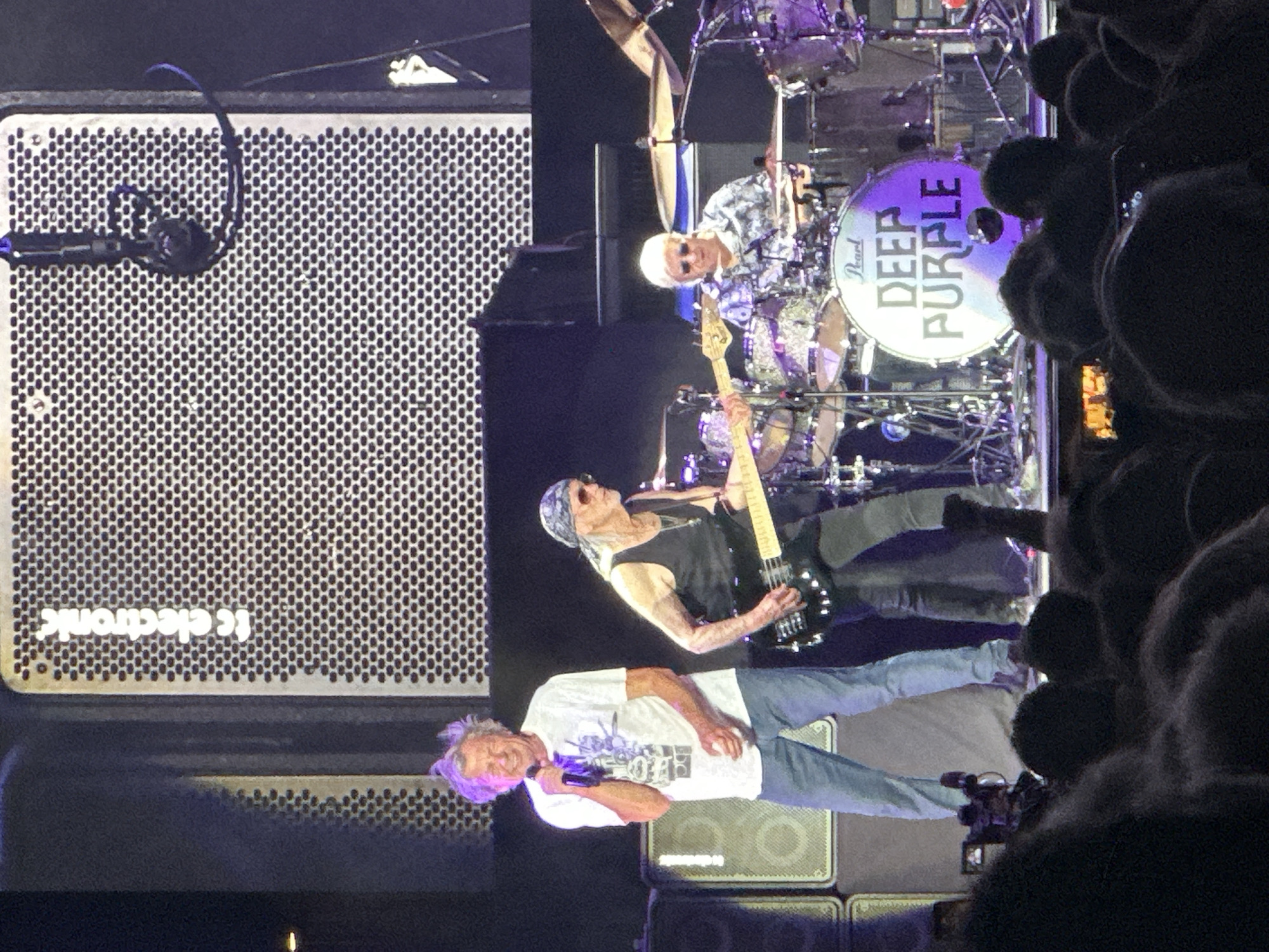 A band named Deep Purple performs on stage in Jerez, Spain. The lead singer, a man with gray, wind-swept hair, is holding a microphone and wearing a white t-shirt and blue jeans. To his right, a man with a bandana and sunglasses is playing a bass guitar, dressed in a black tank top and black pants. Behind them, a drummer with white hair and sunglasses is seated at a drum set, which features a bass drum with "DEEP PURPLE" written on it. The stage is set with amplifiers and lighting equipment. The audience is visible in the foreground, out of focus. The setting is likely an indoor concert venue.