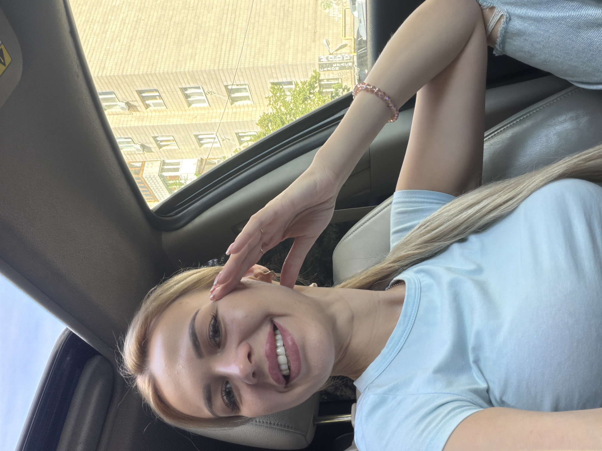 In an outdoor, daytime scene within a car in Kyiv, Ukraine, a fair-skinned woman with blonde hair tied in a ponytail is captured in a selfie. She is wearing a light blue t-shirt and ripped blue jeans. Her right arm is bent, with her hand resting near her face, and she is smiling broadly, revealing her teeth. A delicate pink beaded bracelet adorns her wrist. The car's interior, with its grey leather seats and a sunroof open to the sky, is visible. Through the car window, a multi-story apartment building constructed of light-colored bricks can be seen, with several windows. Some green foliage is also present outside the car. The lighting suggests it is a sunny day.