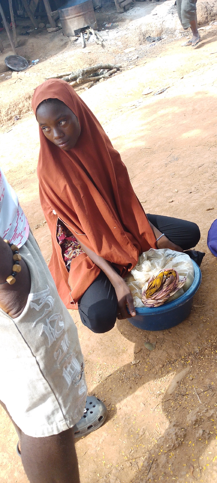 In Gumel, Nigeria, a young individual, likely female based on the attire, is crouched on dusty ground. She is wearing a rust-colored hijab and dark trousers, with a vibrant, patterned fabric visible underneath her hijab. In front of her rests a blue plastic basin containing a white plastic bag. Within the bag, a bundle of colorful fabric, predominantly yellow and pink, is visible. The individual's left hand is resting on the edge of the basin, while her right hand is inside it. The surrounding environment is earthy, with scattered dried leaves and twigs on the ground. In the background, a rudimentary structure constructed from wood and metal is visible, suggesting a rural or market setting. The lighting indicates it is daytime, with strong shadows cast across the scene. Another person's legs, wearing a light-colored garment and dark footwear, are visible to the left of the frame, partially obscuring the main subject.