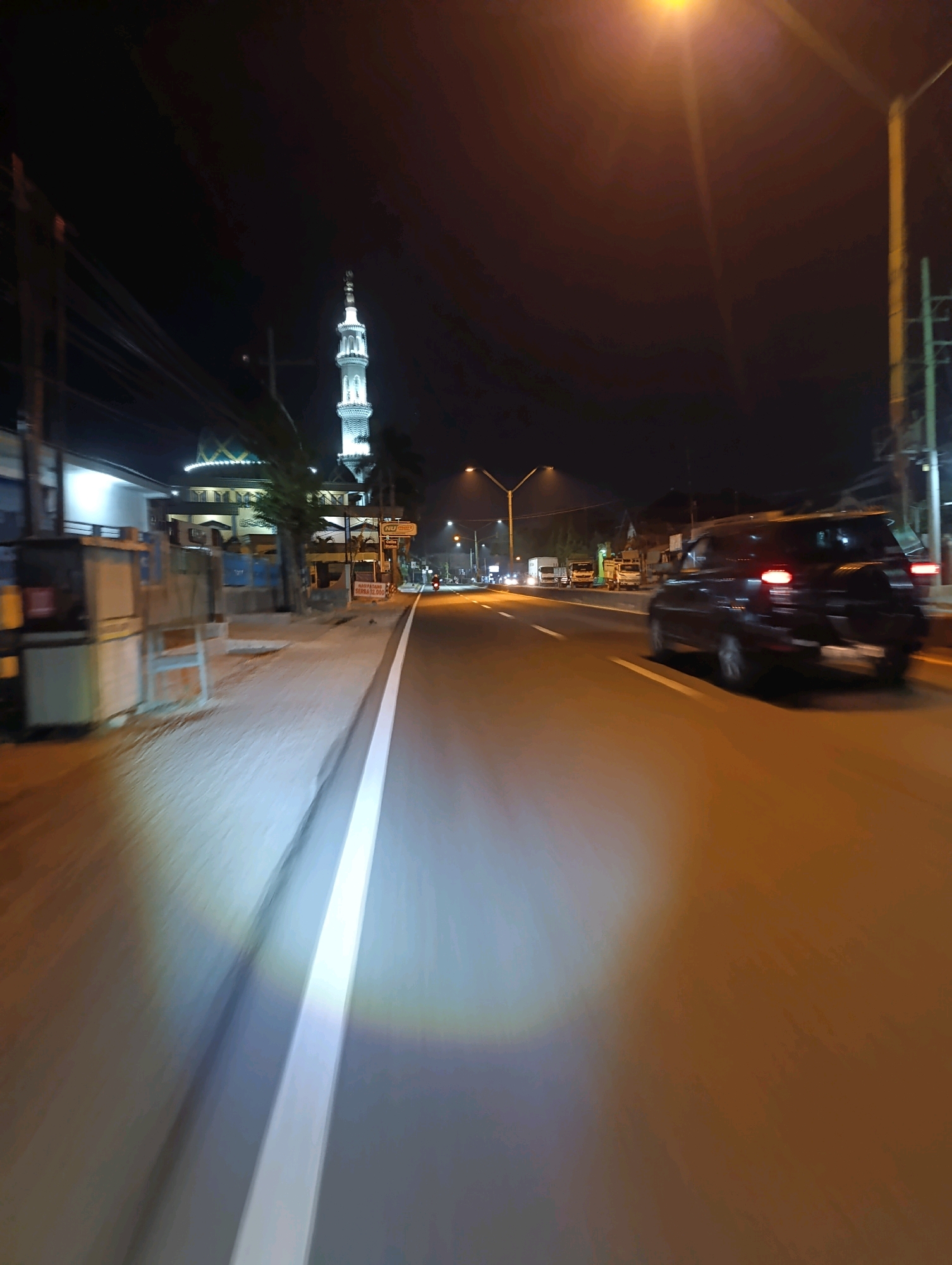 The image depicts a nighttime scene on a street in Glagah Sari, Indonesia. A minaret of a mosque, illuminated with white lights, rises prominently on the left side of the frame, behind a low-rise building with a green, patterned roof. The street is paved and has white lane markings. A dark-colored SUV is in motion on the right side of the road, its taillights illuminated. Several trucks are visible further down the road on the left, also in motion. A streetlamp with a bright orange glow is visible in the upper right portion of the image. The foreground shows a section of the sidewalk with some structures that appear to be kiosks or stalls, and a bright, diffused light source is illuminating the pavement. The image has a motion blur effect, suggesting it was taken from a moving vehicle.