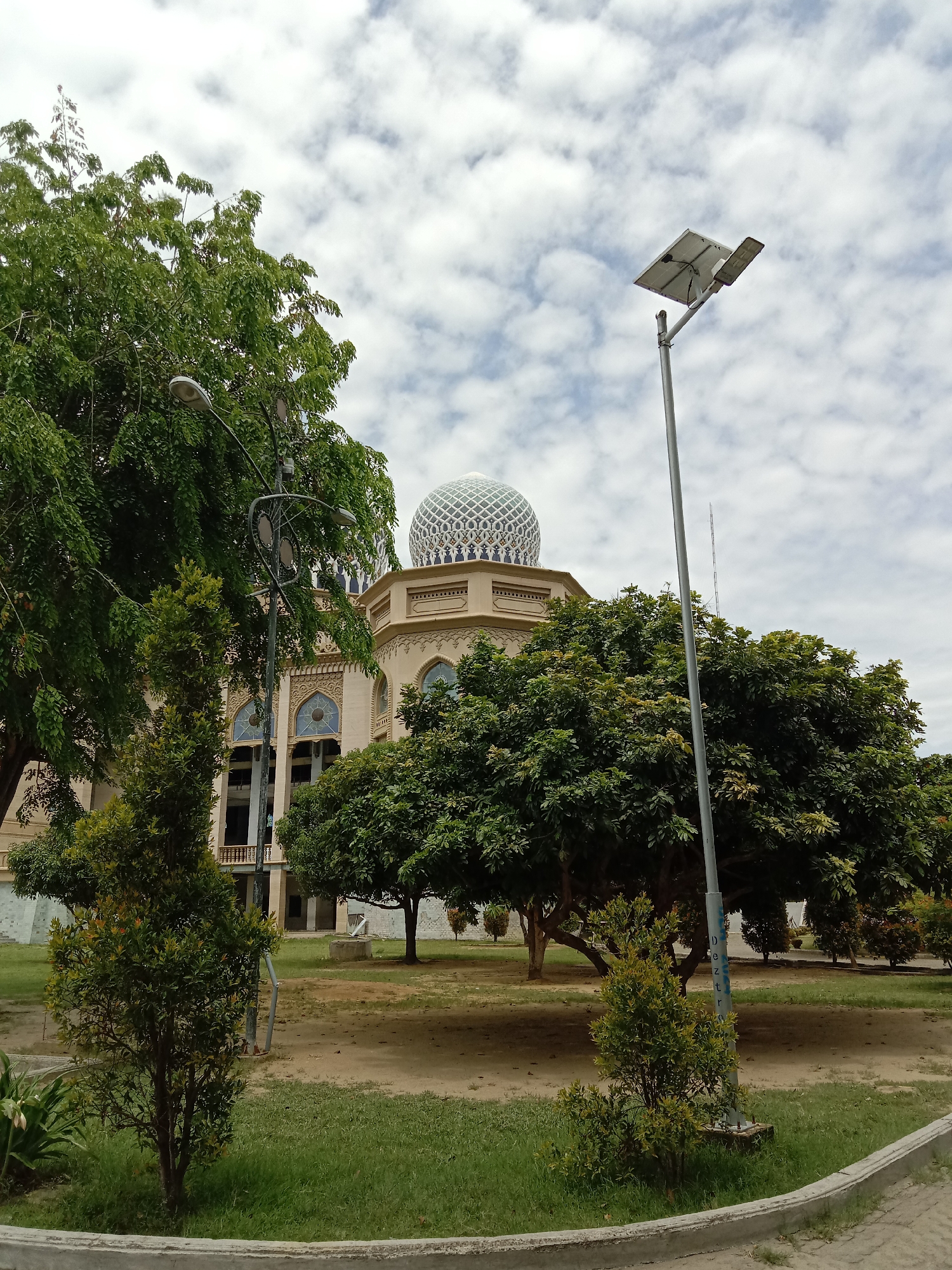 The image shows a mosque with a patterned dome in Lhokseumawe, Indonesia. The building is tan-colored with architectural details above arched windows. Green trees and shrubs partially obscure the view of the mosque. Two types of streetlights are visible: an older style with multiple lamps and a newer solar-powered light. The ground is a mix of patchy grass and dirt. The sky is overcast with clouds.