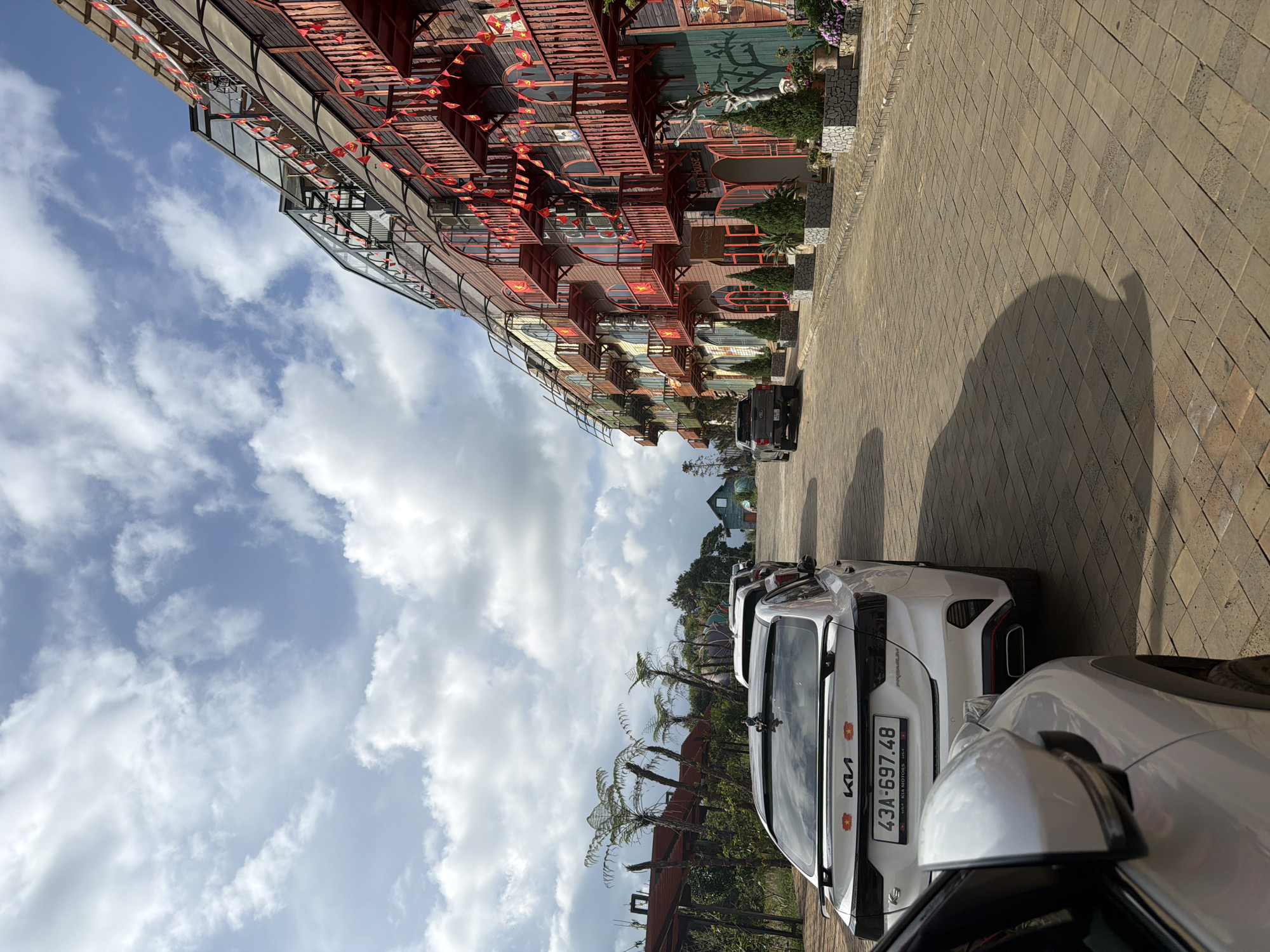 This outdoor image, taken in Măng Đen, Vietnam, features a distinctive multi-story building and several parked cars under a bright, partly cloudy sky. The building boasts unique and intricate architecture, characterized by numerous red-orange balconies and railings, glass elements, and a complex, layered design. Small red and yellow Vietnamese flags are strung decoratively along the building's facade and balconies, while planters with greenery and flowers add to its visual appeal.
A paved road or parking area occupies the foreground and mid-ground, where several cars are parked. The closest vehicle is a white Kia, with "KIA" and "PREMIUM" visible on its rear. Its license plate clearly displays "43A-697.48," along with smaller text "THACO KIA MOTORS" and a small Vietnamese flag symbol. Other cars, including a dark SUV, are parked further down the paved area. The ground is paved with rectangular blocks, and the presence of clear shadows indicates bright daylight. Palm-like trees or ferns are visible in the landscape around the building. The sky is a clear blue with scattered white clouds, suggesting pleasant weather. The scene portrays a unique architectural setting, likely a hotel or resort.
