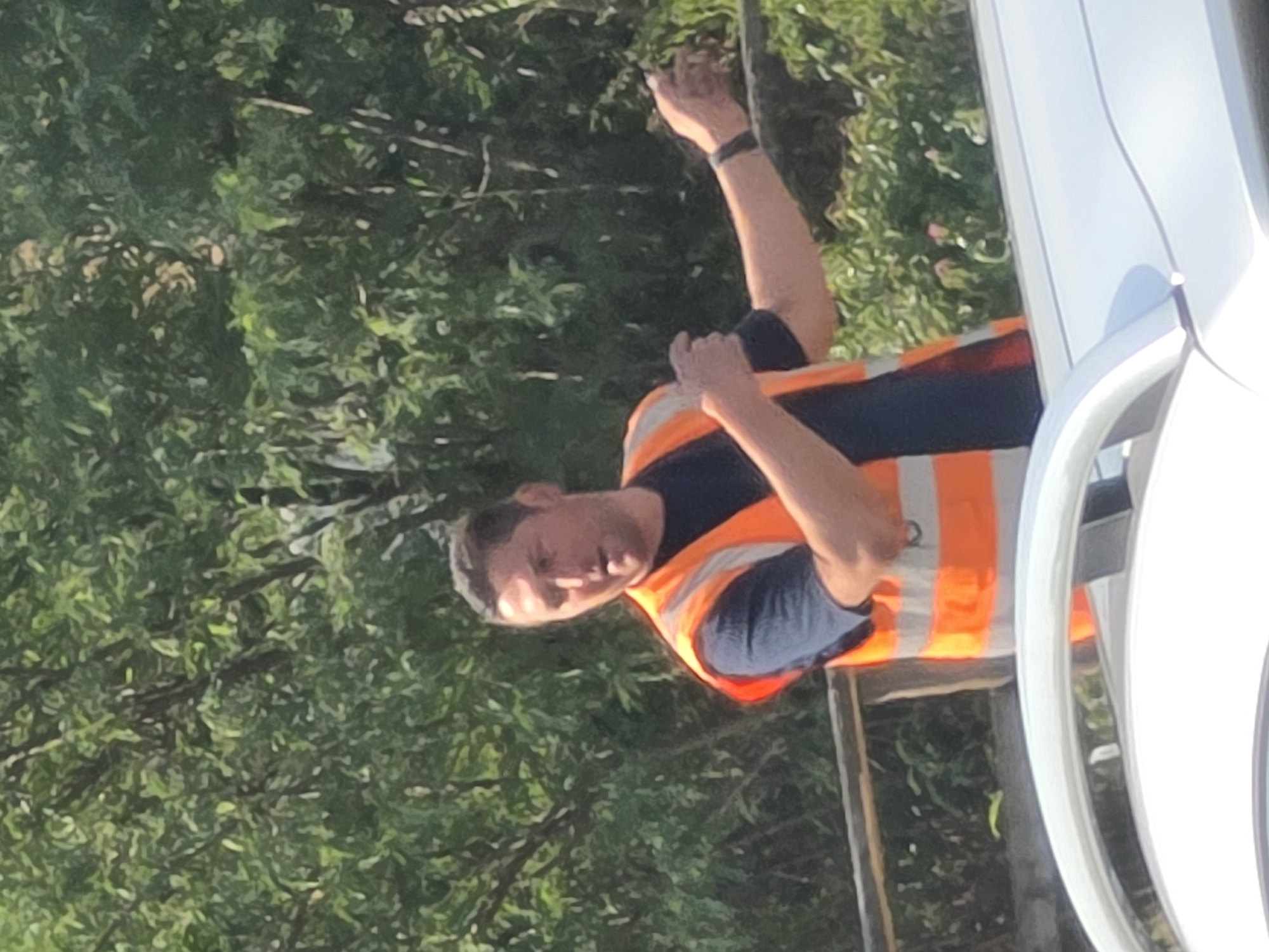 A man wearing an orange reflective vest over a dark shirt stands outdoors in front of dense green foliage. His right arm is extended, with his hand open and palm facing forward. He is looking to his right and appears to be speaking. A dark band is visible on his left wrist. The lower portion of the image shows the curved, reflective surface of a car. A wooden railing is present in the midground. The image does not contain information that places it in Frankfurt, Germany.
