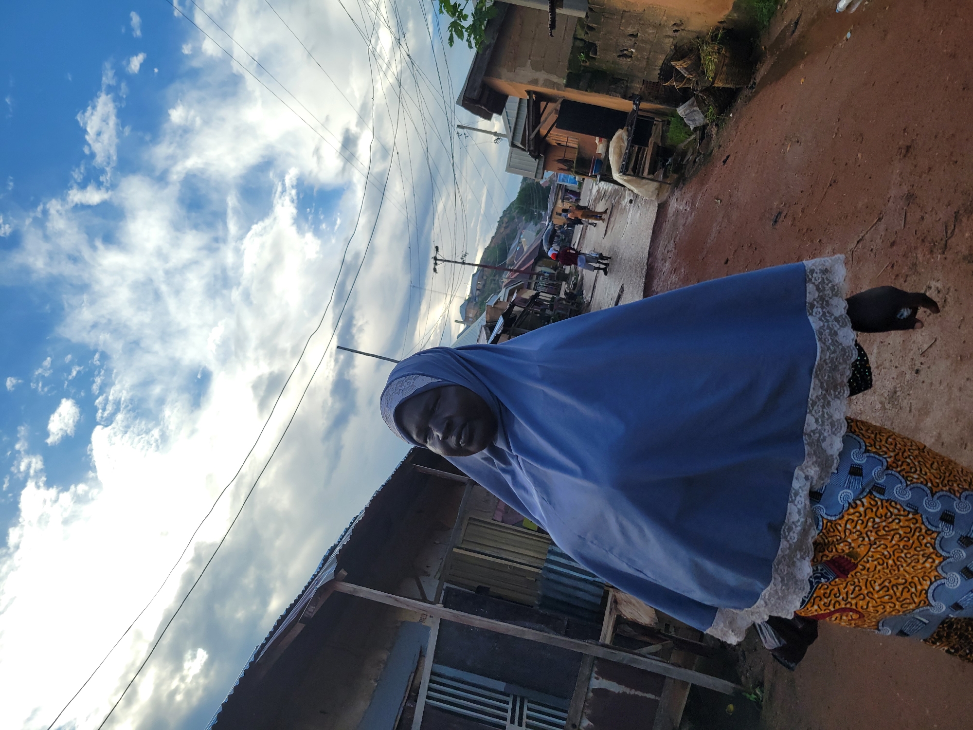 A full shot shows a Black woman in a blue hijab and an orange and blue patterned skirt walking down a dirt road in what appears to be a village. The woman is looking to her right, and her hands are at her sides. The road is wet, suggesting it has recently rained. In the background, there are several small buildings, some with corrugated metal roofs. A few other people are visible in the distance, walking down the road. The sky is cloudy with patches of blue.