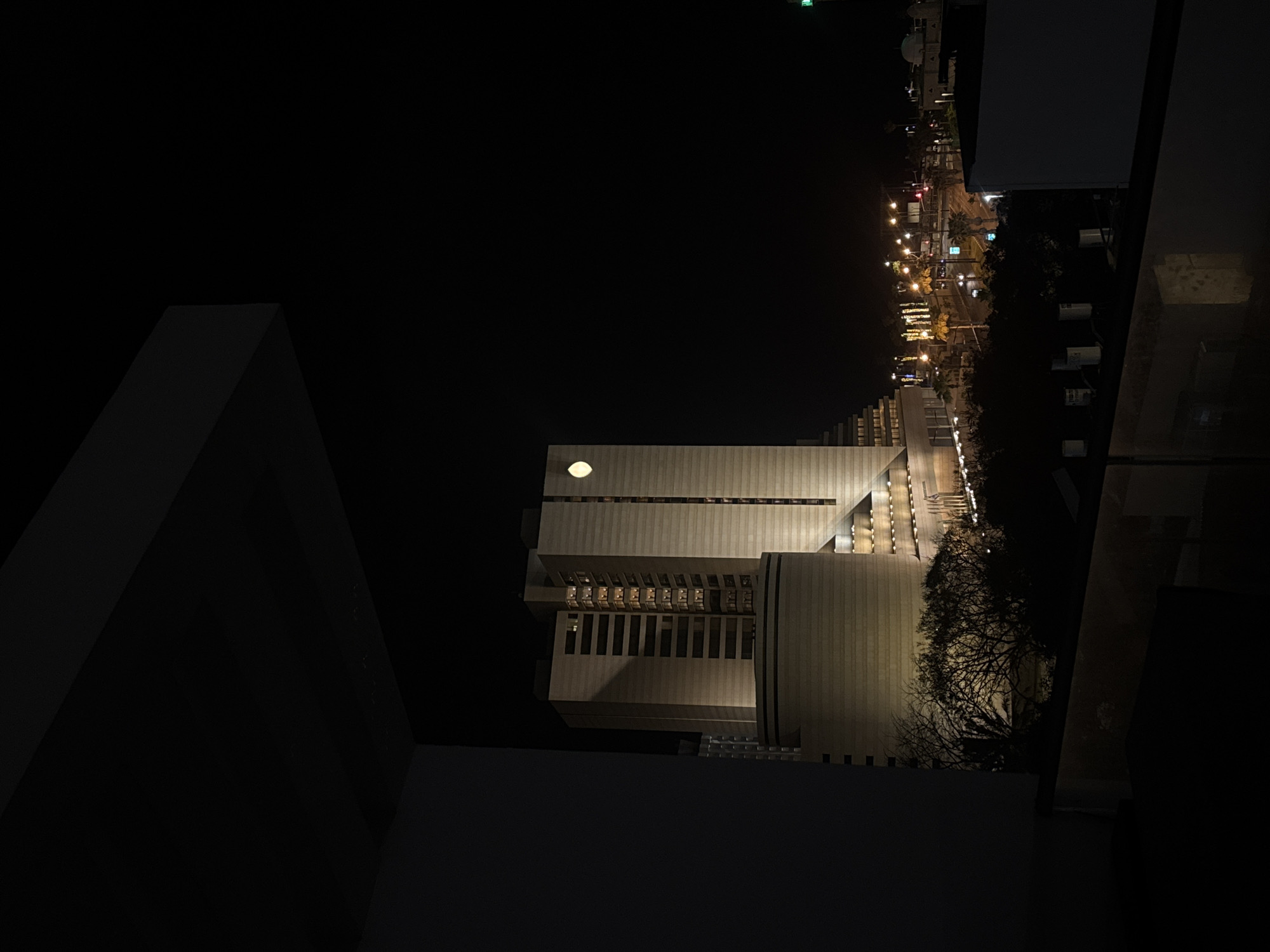 The image depicts an aerial nighttime view overlooking Tel Aviv, Israel. Predominantly dark, the scene is illuminated by artificial lighting. A modernist building with illuminated windows and a prominent circular light fixture is the focal point. Adjacent to the building, a street is lined with illuminated streetlights and possibly some buildings. Trees are also visible along the street. The foreground is dominated by architectural elements, likely part of the structure from which the photo was taken.