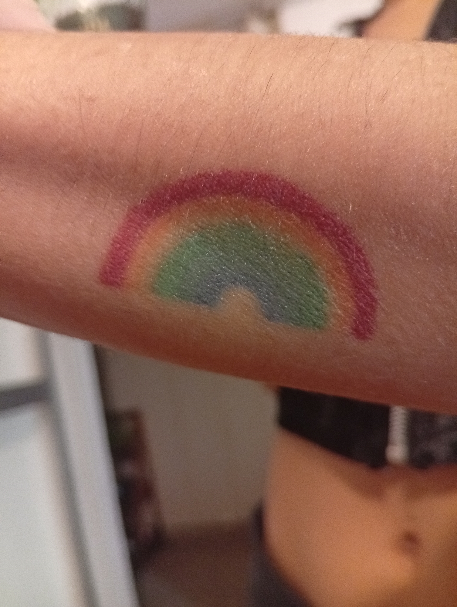 A close-up shot depicts a forearm with a rudimentary rainbow painted on the skin. The rainbow is semi-circular, comprising red, green, and blue hues. The skin is pale with fine hairs. In the background, partially visible, are elements suggestive of a domestic setting, including a blurred white object (possibly a refrigerator) and a person wearing a black top with a zipper. The overall scene conveys an informal and personal context. There is no location data present in the image. The city of Buenos Aires, Argentina is not evident in the image.