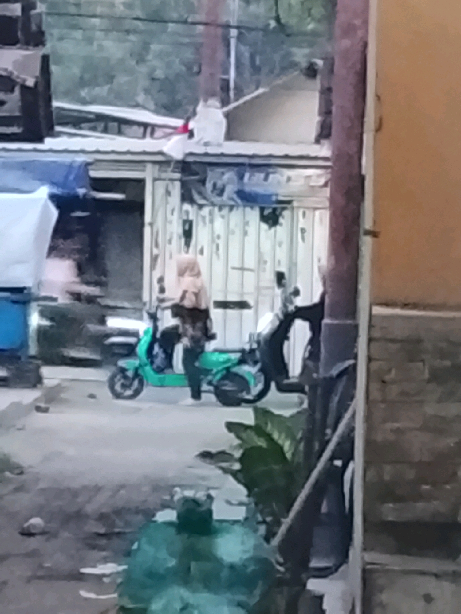 The image depicts an outdoor scene in Ujungrusi, Indonesia. A person wearing a light brown hijab and a dark patterned top is standing next to a teal-colored scooter. They are holding onto the scooter's handlebars. Behind the scooter, another person is partially visible, appearing to be seated on a black scooter. The background consists of simple buildings with corrugated metal roofs and siding, some with open doorways. Vegetation, including a large leafy plant, is visible in the foreground and midground. The ground is paved and appears somewhat rough. The overall lighting suggests it is daytime, possibly overcast.