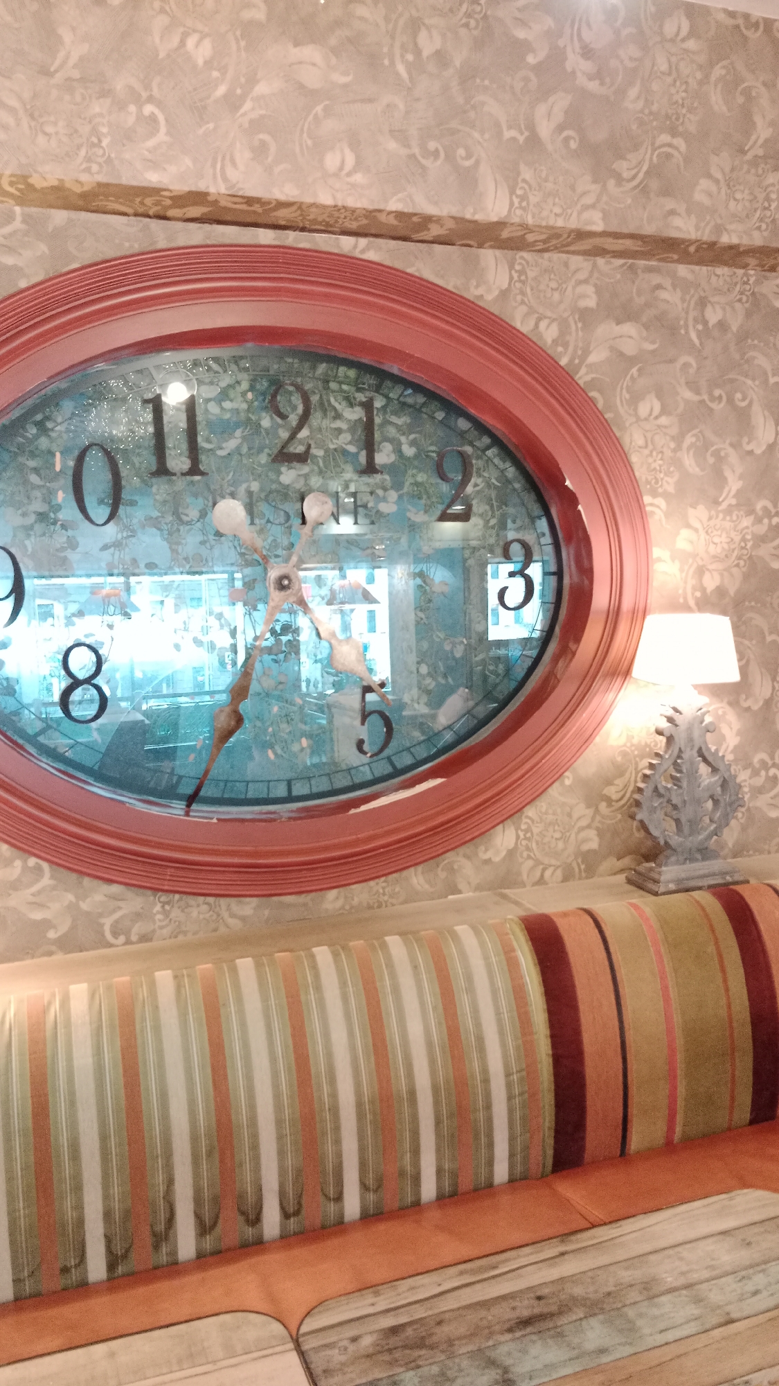 Here is a detailed description of the image:
The image depicts an interior setting in the city of Unknown, Unknown. Dominating the frame is a large, oval-shaped clock with a coral-colored frame. The clock face features black numerals and decorative hands, set against a patterned background that suggests floral or leafy motifs. The clock's face is somewhat reflective, showing a glimpse of the surroundings beyond.
Below the clock is a striped sofa with varying colors including orange, green, and maroon. The stripes are vertical and of alternating widths. The sofa appears to have a wooden base with a distressed or weathered finish.
To the right of the clock, positioned on a surface near the sofa, is a table lamp. The lamp has a white lampshade and an ornate, gray-toned base.
The background wall is covered with a patterned wallpaper, featuring a subtle, swirling design in light, muted tones.
The lighting in the scene appears to be a mix of natural and artificial, with the lamp contributing to the overall illumination. The setting suggests a waiting area or a lounge space, possibly in a hotel or a public building.