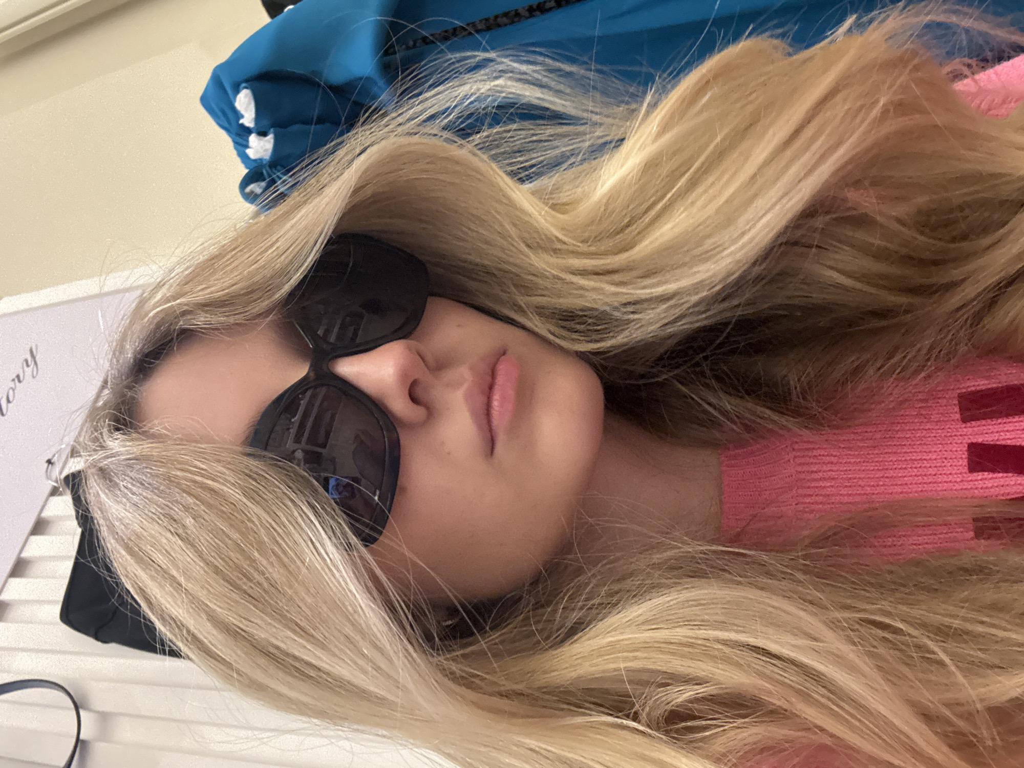 A blonde woman wearing sunglasses and a pink sweater lies down. A blue garment hangs above her. A white surface with lettering is visible. The location is unknown, but may be Mexico City, Mexico.