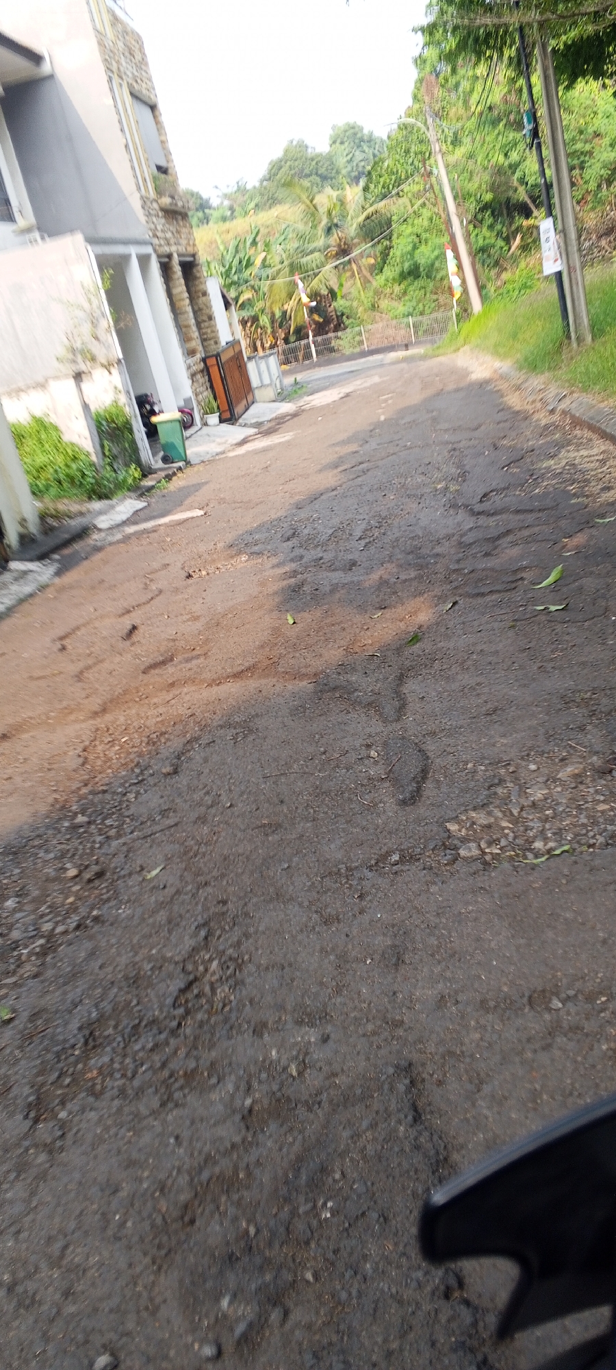 The image shows a street in Gunung Putri, Indonesia, with a modern building on the left and a lush, green hillside with palm trees in the background. The road itself is paved with asphalt and appears to be in disrepair, with several potholes visible. There is a green trash bin near the building. A motorcycle is partially visible in the foreground, suggesting it is the viewpoint. The lighting suggests it is daytime, with sunlight casting shadows on the road. The overall impression is of a somewhat neglected but otherwise scenic urban environment.