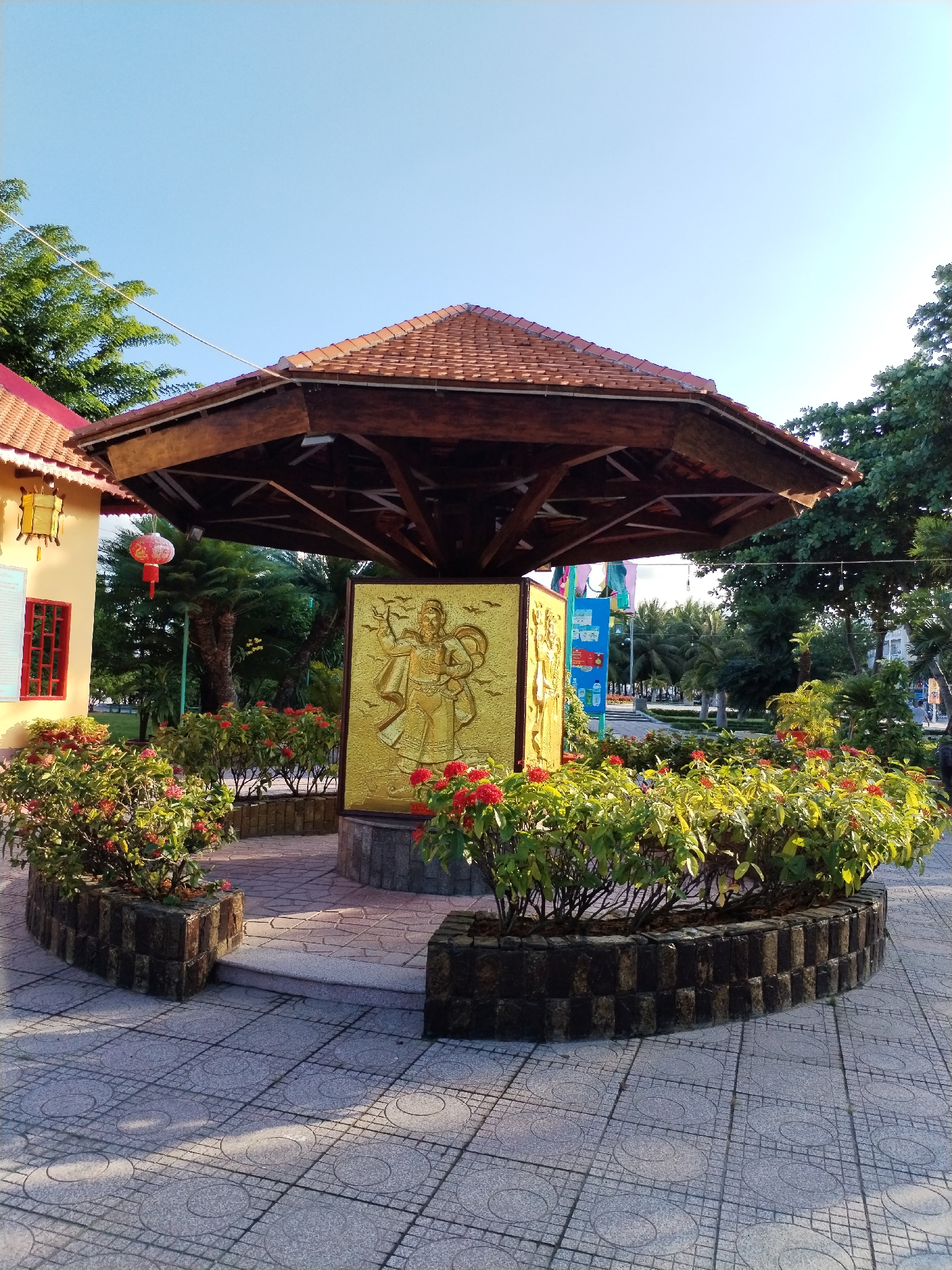 The image depicts an outdoor scene in Bac Nha Trang Ward, Vietnam. A centrally located, hexagonal-shaped structure with a brown tile roof and dark wood supports stands prominently. The structure features a gold-colored relief artwork on its sides, depicting a figure in traditional attire. The artwork is surrounded by bushes with red flowers. The structure is situated on a raised platform with a step leading up to it, and the ground is paved with gray tiles featuring a circular pattern. To the left of the structure, a small, yellow building with a red-framed window and a red lantern is visible. Trees and other greenery surround the area. The sky is clear and blue.