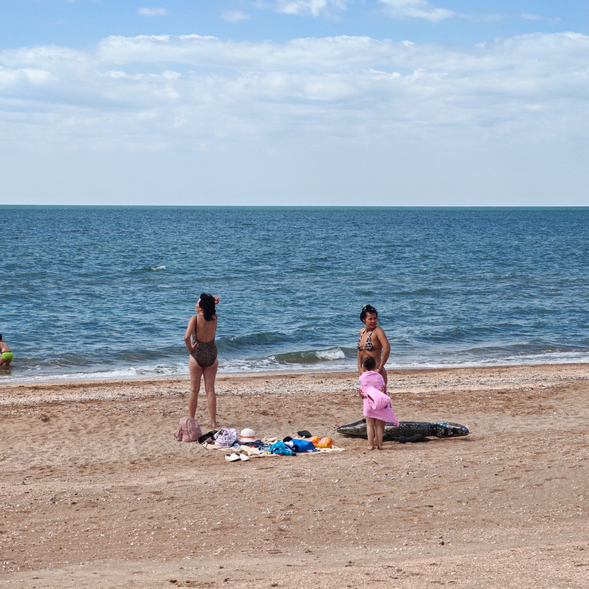 The image shows a beach scene in городское поселение Щелкино, Ukraine. Two women in swimsuits stand on the sand near the water's edge. A child in a pink towel stands near one of the women. A blanket with various items including clothes, hats, and shoes lies on the sand. Near the blanket is an inflatable crocodile. The sky is blue with scattered clouds. The water is blue-green.