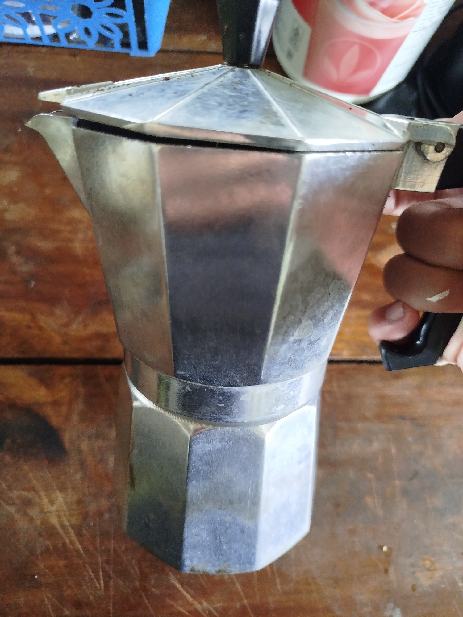 An overhead shot shows a used aluminum Moka pot on a weathered wooden surface, likely a table, in Dukupuntang, Indonesia. The Moka pot is octagonal and appears to be in the process of cooling down, with a hand holding its black handle. A blue plastic container is visible in the upper left corner, and a portion of a pink and white beverage container is in the upper right. The wooden surface is marked with scratches and a few dark stains, suggesting outdoor or heavily used conditions.