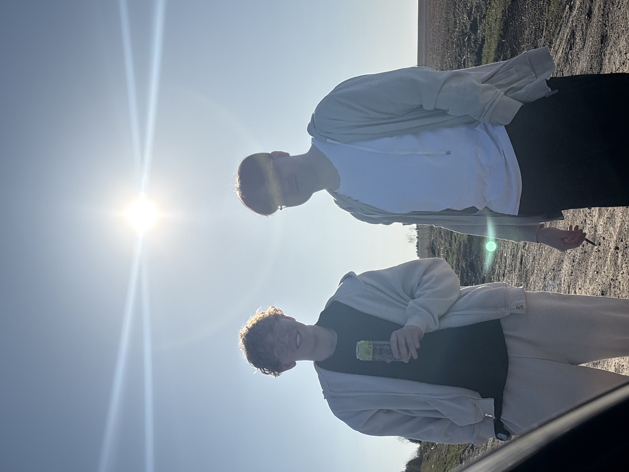 Two individuals stand outside on a sunny day. One person is wearing a white t-shirt and a light-colored jacket, while the other person is wearing a dark t-shirt and a light-colored jacket. One of them is holding a beverage. They are likely standing in an open area, possibly a field or parking lot. The location is Баклуши, Russia.