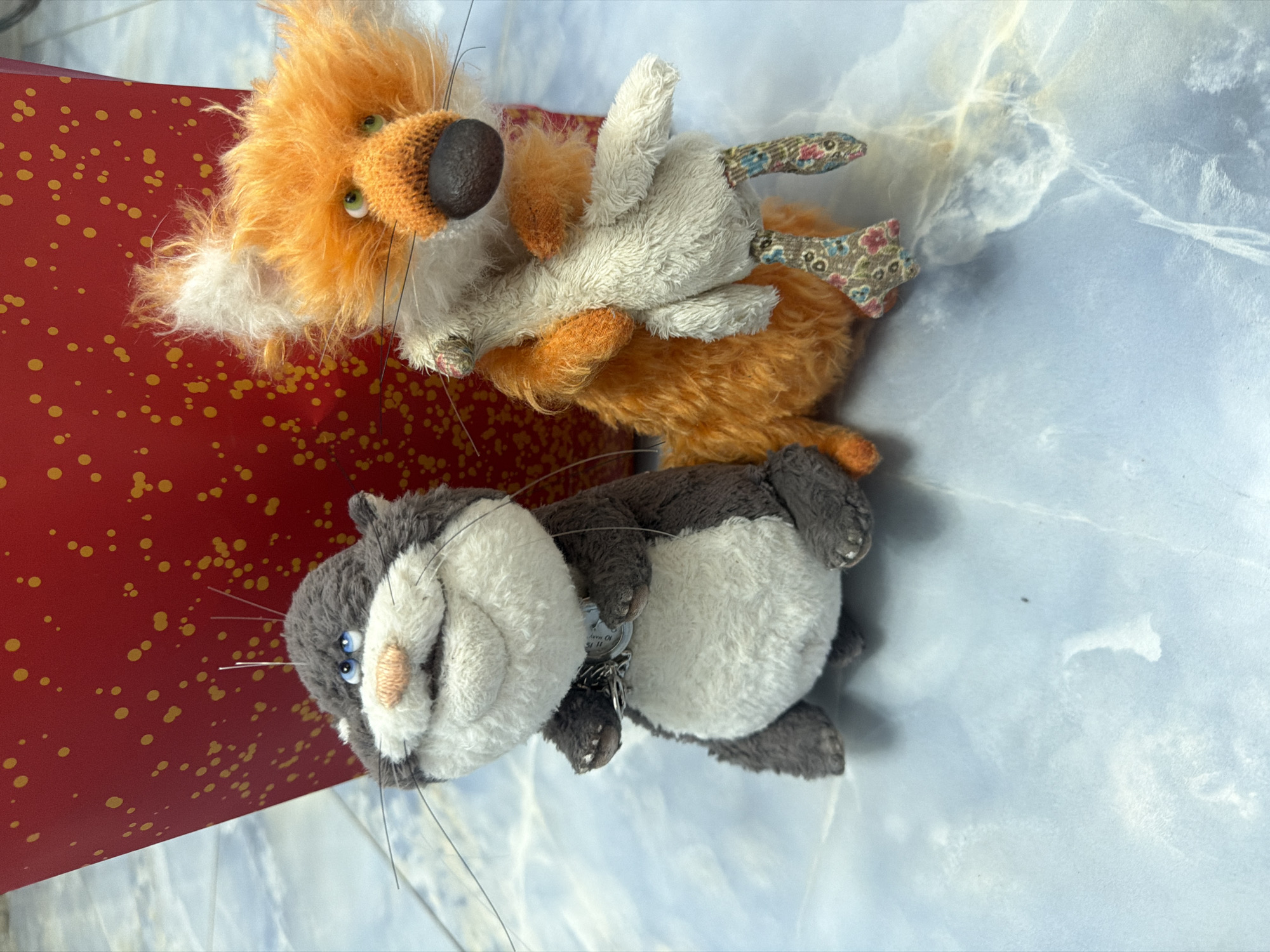 Two stuffed toys, a grey cat and an orange fox, are positioned on a light blue tiled surface. The cat, on the left, has blue eyes, a pink nose, and a silver tag around its neck. The fox, on the right, is holding a white stuffed toy and has green eyes and an orange knitted snout. Behind the toys, a large red object with yellow speckles is partially visible. The lighting suggests an indoor setting.