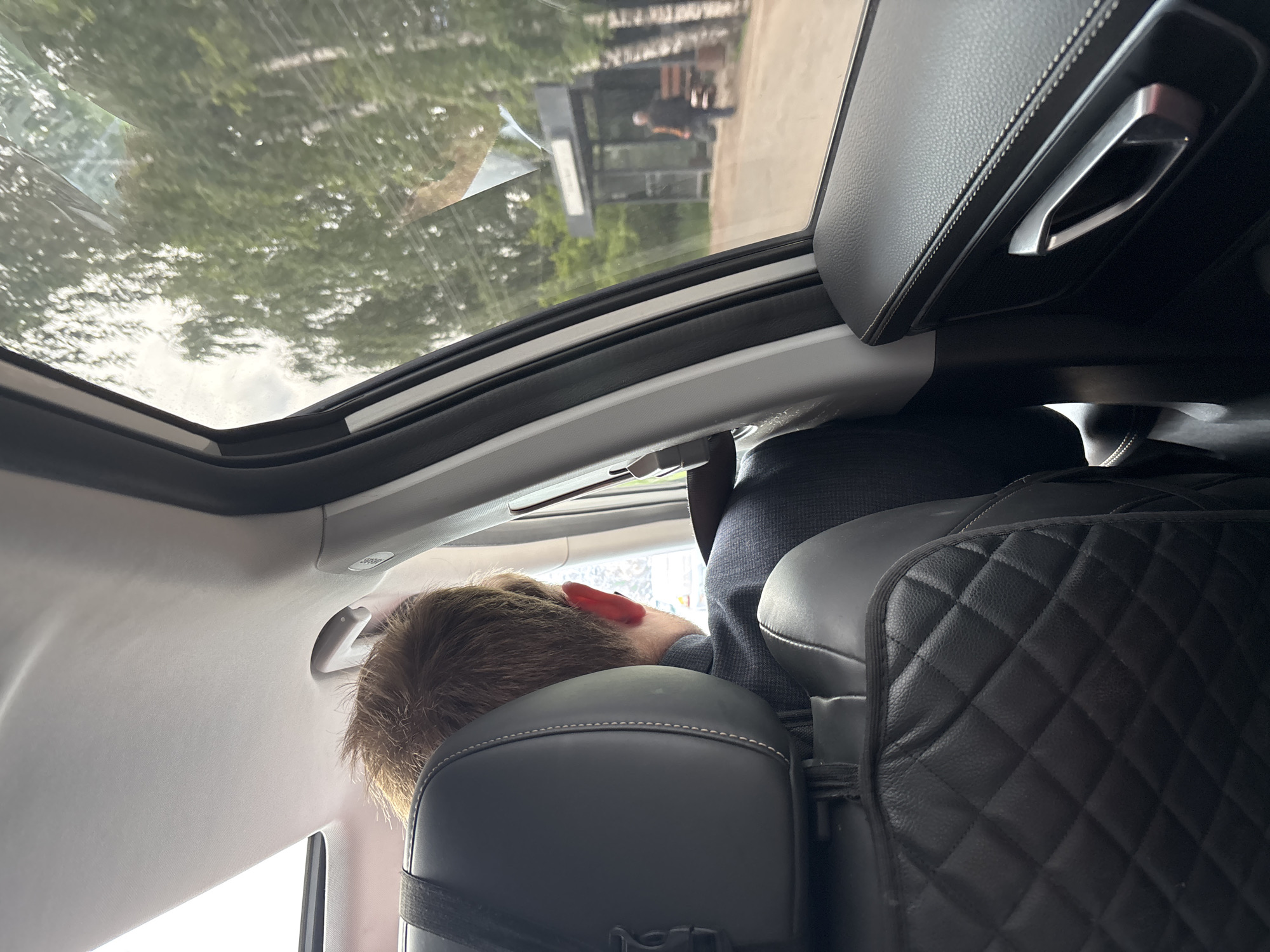 The image shows the interior of a car, looking out the window to a street scene in Chaikovsky, Russia. A person's head and shoulders are visible in the foreground, looking out the window. Outside, there is a bus stop with a person standing near it. Trees and buildings are visible in the background. The car interior appears to be modern and well-maintained, with leather seats and a sunroof. The overall lighting suggests it is daytime.