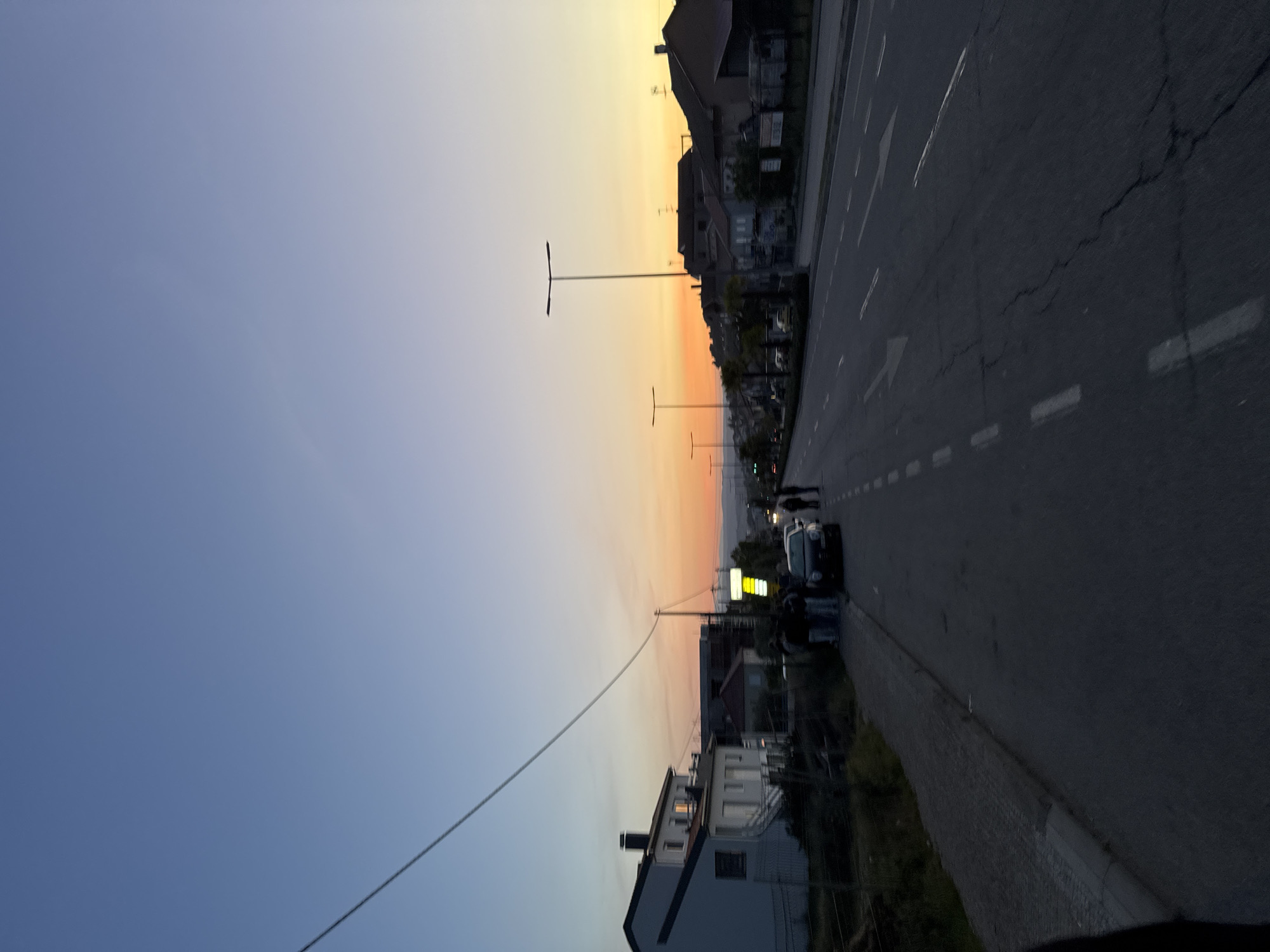 The image depicts an urban setting in Bragança, Portugal, during dusk. The sky transitions from blue to orange-yellow. A main street with asphalt pavement and lane markings runs parallel to buildings on the right. Lampposts line the street. A group of people stands near a parked car on the side of the road. Power lines stretch across the sky.