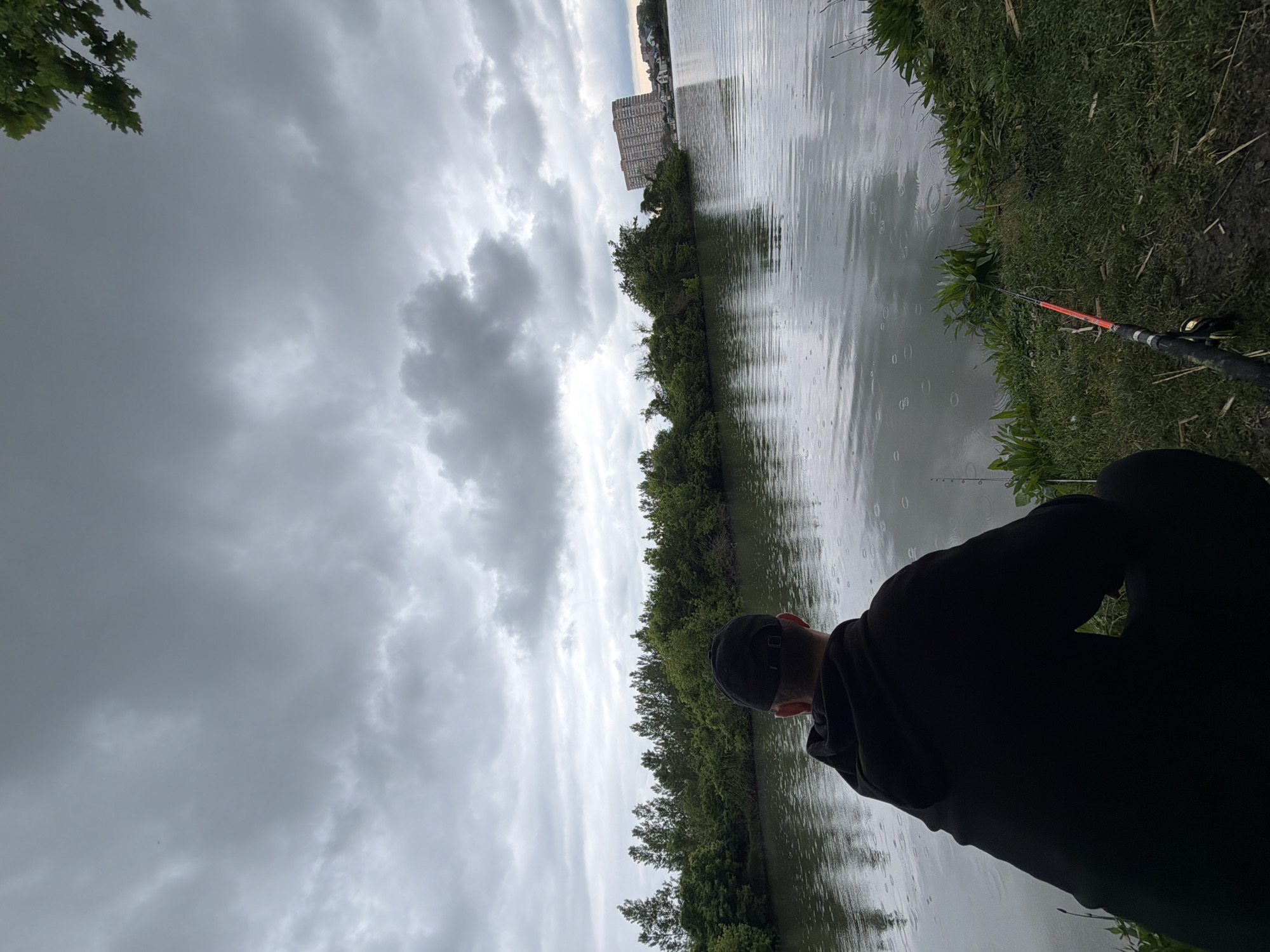 The image shows a person fishing by a lake in Krasnodar, Russia. The person is wearing a black hoodie and a cap, facing the water. A fishing rod is visible near the edge of the bank. Rain is falling, creating ripples on the lake's surface. Trees line the opposite shore, with buildings visible in the distance. The sky is overcast with grey clouds.