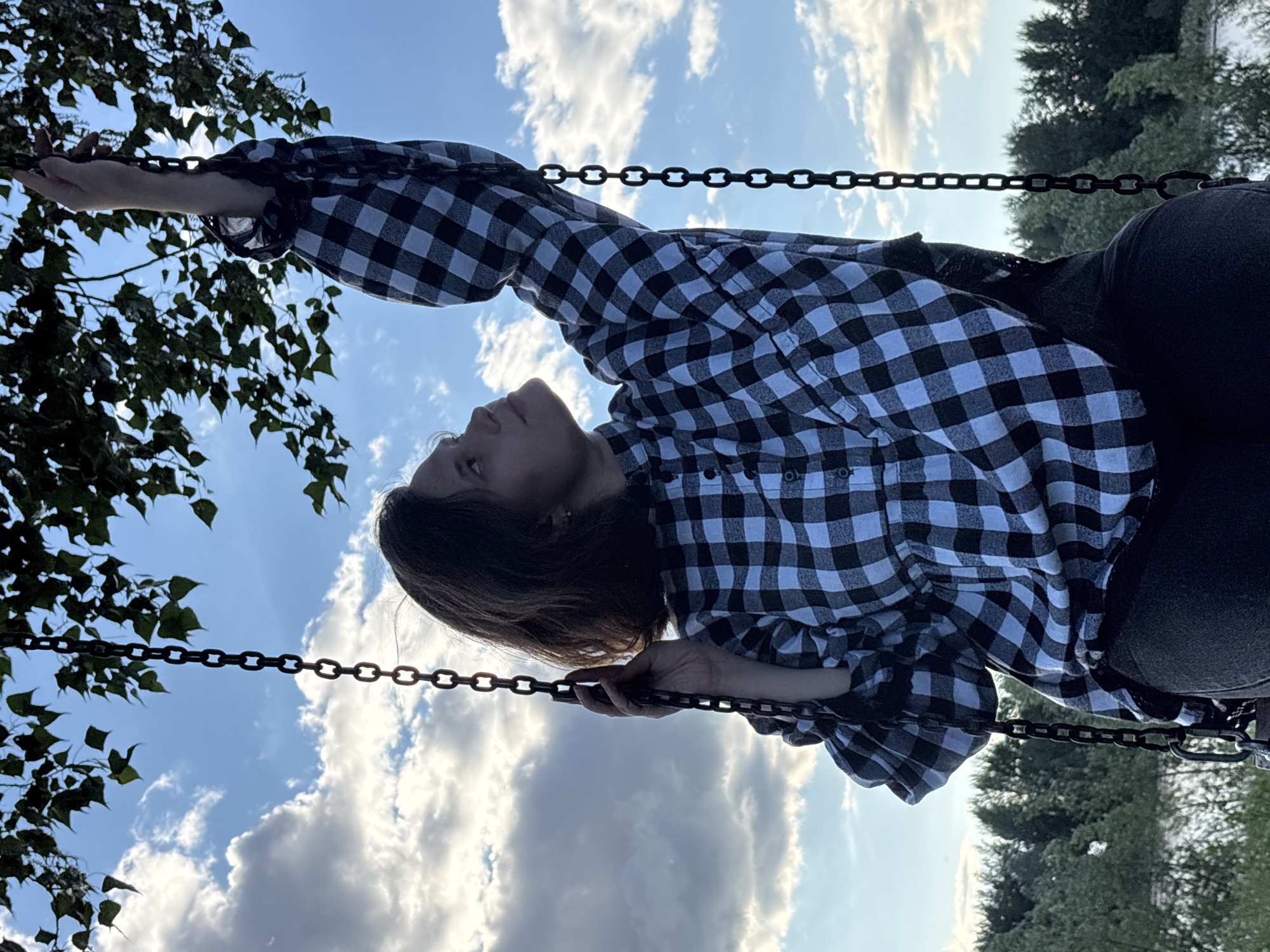 A young woman is seated on a swing, looking upwards and reaching for a branch. She is wearing a black and white checkered shirt and dark pants. The swing is suspended from a tree by thick chains. The background features a cloudy blue sky, lush green trees, and a body of water. The lighting suggests it is either early morning or late afternoon. The location is identified as Unknown, Unknown.