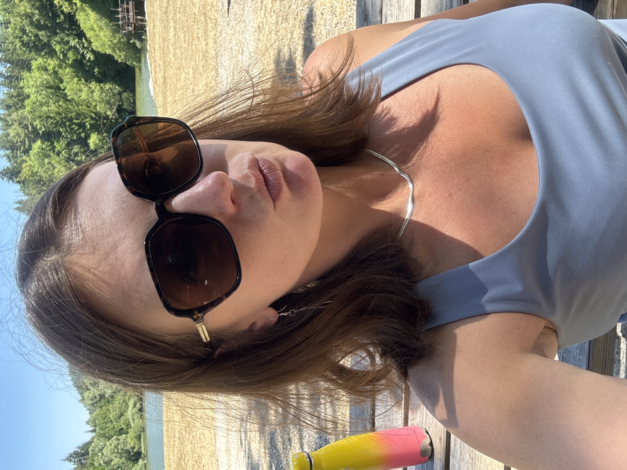 A woman is taking a selfie on a sunny day, seated on a wooden bench in what appears to be a park or outdoor recreation area. She is wearing sunglasses, a grey tank top, and a silver necklace. A yellow and pink water bottle is positioned to her left. In the background, there is a body of water, sand, trees, and a small wooden structure. The location is identified as Coteaux du Lizon, France.