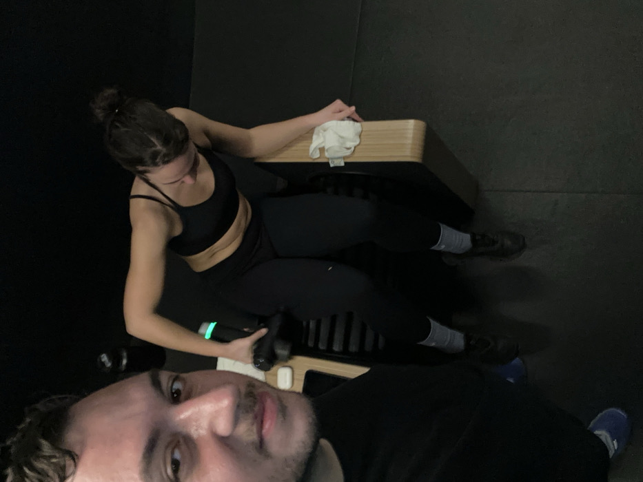 A male subject, partially visible on the left foreground, is capturing the scene with a camera. He has dark hair, a slight mustache, and wears a dark top and a blue shoe on his left foot. A white AirPods case rests on a light wooden surface near him. In the mid-ground, a female subject with dark hair tied in a bun is seated on a smart strength exercise machine, characterized by a light wooden top edge and a dark, ribbed base. She wears a black sports bra, black leggings, grey socks, and black athletic shoes. Her right hand operates a black percussion massager, emitting a green light, which she applies to her upper left thigh. A white towel is folded on the wooden edge of the machine near her left hand. The background features dark walls and a dark rubber floor, typical of a high-end fitness studio. A dark water bottle is visible in the background. This indoor fitness facility is located in the city of London, United Kingdom.
