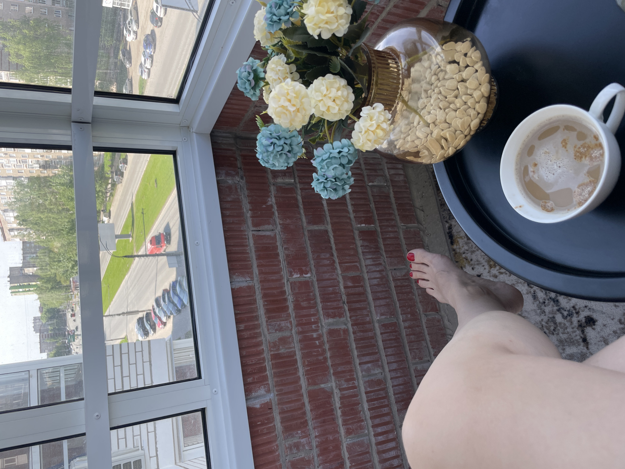 A top-down shot captures a scene on a balcony. A woman's leg and foot with red nail polish are visible. Adjacent is a small round table made of black material, holding a white coffee mug and a floral arrangement in an amber vase filled with beige stones. A brick wall extends upwards, meeting a window frame with a view of a street with parked cars and distant buildings. The location is an unidentified urban area.
