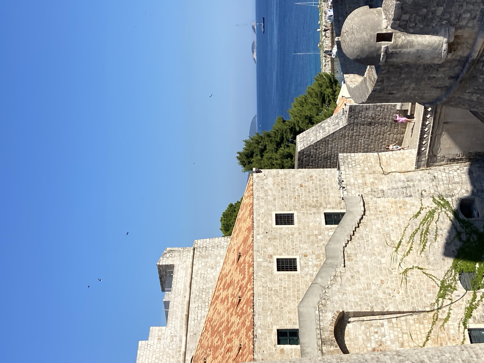 The image shows a view of the walls of Dubrovnik, Croatia, on a clear, sunny day. The foreground is dominated by the stone fortifications of the Old Town, featuring weathered stone blocks, arched doorways, and a tiled roof on one of the buildings. A staircase winds upwards along the wall, and some greenery is climbing up the stonework. Two women are visible walking on a walkway along the top of the fortifications. In the background, the deep blue Adriatic Sea stretches out, dotted with a few islands and a sailboat. The sky is a bright, cloudless blue with a few birds in flight. The scene evokes a sense of history and a Mediterranean coastal atmosphere.
