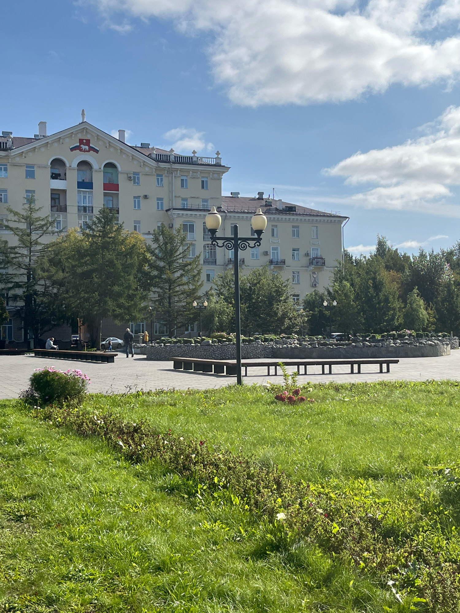 The image depicts a view of a park or public square in Perm, Russia, on a sunny day with scattered clouds. In the background, a light-yellow, multi-story building with a distinctive architectural style, featuring a pediment with a crest and what appears to be a Russian flag motif, dominates the scene. The building has numerous windows and balconies. In front of the building, a line of mature evergreen trees stands, casting shadows.
The mid-ground features a paved area with several dark wooden benches arranged around a circular, low-walled structure filled with stones and some foliage. A black lamppost with a yellow light fixture stands prominently near the left side of this paved area. A few people are visible: one person is sitting on a bench on the left, and two figures are walking in the central paved area. A light-colored car is parked further back.
The foreground consists of a well-maintained grassy area with some flowering bushes, characterized by green grass and scattered pink and red flowers. The overall impression is one of a pleasant urban outdoor space.