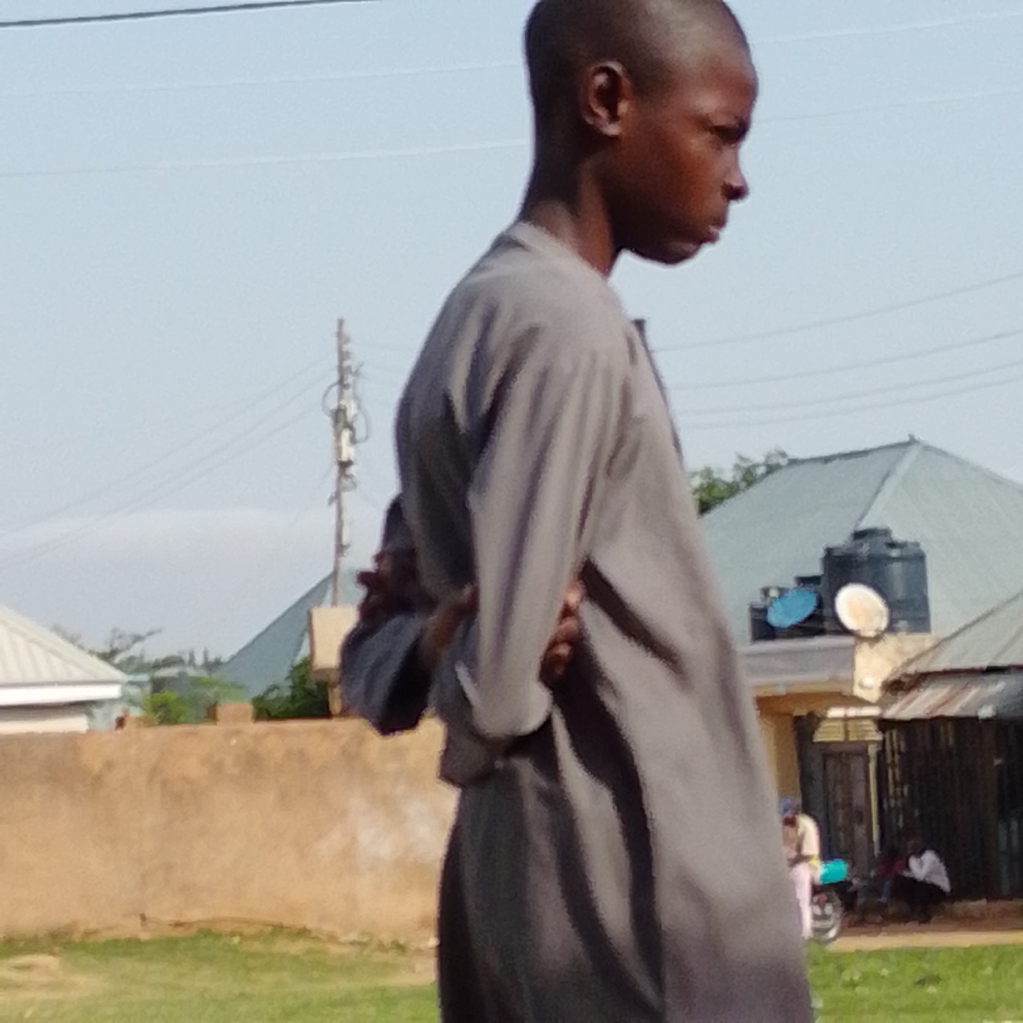 The image depicts an outdoor scene in Gombe, Nigeria. A young male, with a closely cropped haircut and dark skin tone, is prominently featured in the foreground, facing right and looking downwards. He is wearing a grey, loose-fitting garment that covers his torso and extends to his legs. His right arm is bent, with his hand resting on his back, while his left arm is obscured by the garment.
In the background, various elements of a semi-urban or rural environment are visible. A beige-colored wall, likely a boundary, runs horizontally across the middle of the frame. Behind the wall, structures with corrugated metal roofs and lighter-colored walls are present. A utility pole with wires is situated to the left of the main subject. Further back, a water tank on a raised platform, possibly a water harvesting system, is visible on top of a building, alongside what appears to be satellite dishes. In the far background, a green, sloped landscape suggests hills or mountains under a hazy sky.
Several other individuals are present in the background, though their details are less clear due to distance and the overall image quality. To the right of the water tank, a person in a light-colored garment is standing near a motorcycle. Further to the right, in front of a dark, shed-like structure, at least two individuals are discernible, one of whom appears to be wearing a white shirt and dark pants, and is crouching. The ground in the foreground is covered in dry grass.