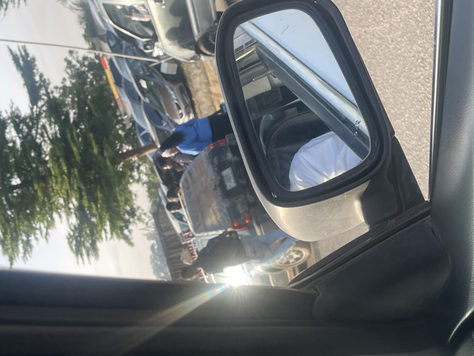 The image, taken from inside a vehicle, shows a traffic jam in Dutsen Alhaji, Nigeria. The side mirror of the car captures a reflection of the surrounding scene: several cars are lined up, with people standing outside their vehicles, some wearing casual clothing. The background features trees and other vehicles. The presence of people outside their cars, combined with the congested traffic, suggests a possible disruption or accident on the road. Sunlight is visible in the distance, creating lens flare.