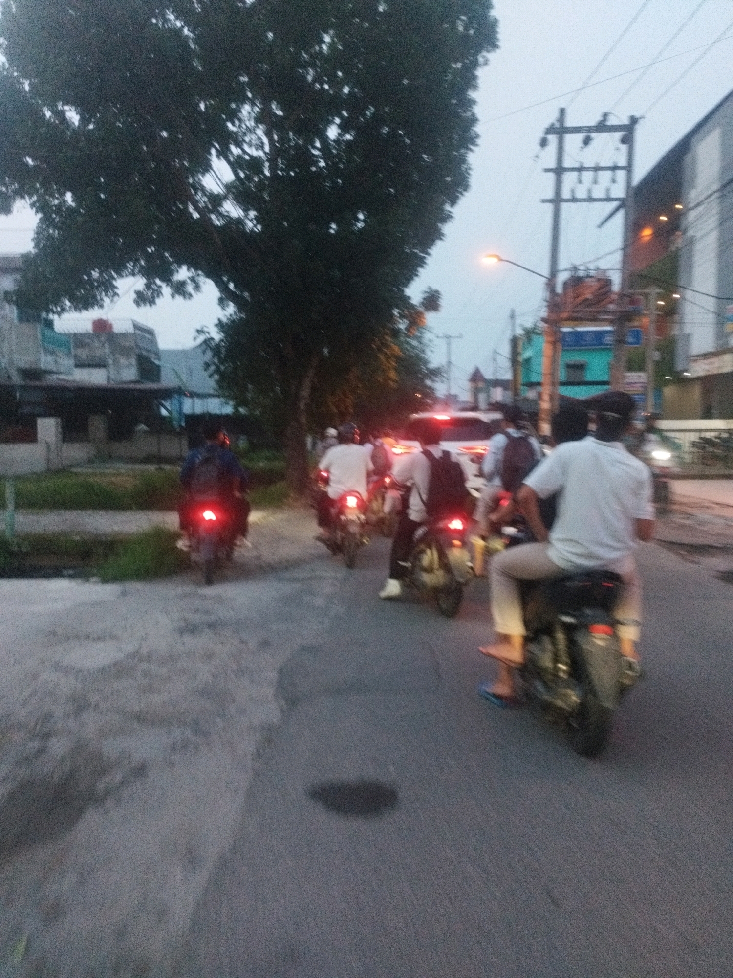An outdoor, low-light, long-shot, full-frame photograph taken in the City of Medan, Indonesia, captures a street scene with multiple motorcycles. In the foreground, a person on a motorcycle is visible from the rear, wearing a light-colored short-sleeved shirt and light-colored pants, with one foot wearing a blue flip-flop resting on the pavement. The motorcycle has a visible taillight. To the left and slightly behind, another motorcycle with a rider is also depicted from the rear, with a bright red taillight illuminated. Further back, several other motorcycles and riders are seen, some with illuminated taillights, moving along a paved road. Buildings and dense foliage from a large tree are present in the background on the left side of the frame. Overhead, electrical poles and wires are visible against a dusky sky. The overall impression is of a street during twilight or early evening, with traffic on the road.