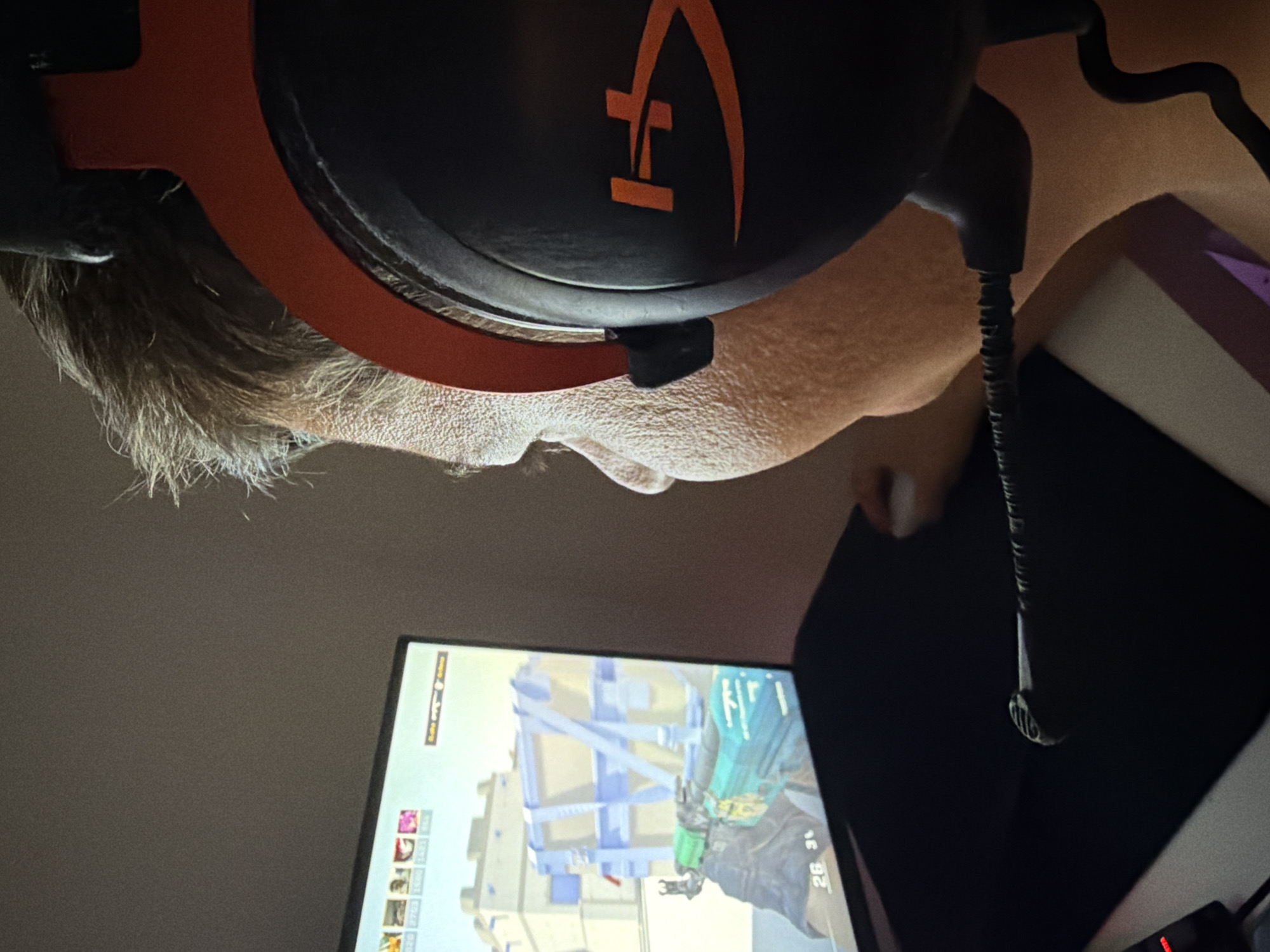An angled close-up shot shows a person wearing a black headset with an orange headband and the logo in orange on the black earcups. The person has short, gray hair and is facing forward toward a computer screen that is visible at the bottom of the frame. The screen displays a video game with a first-person perspective, featuring a structure with blue and white elements. A black mousepad is partially visible to the right of the screen, with the coiled cable of the headset lying on it. The environment is dimly lit. The location is not identifiable as Budva, Montenegro from the available visual information.