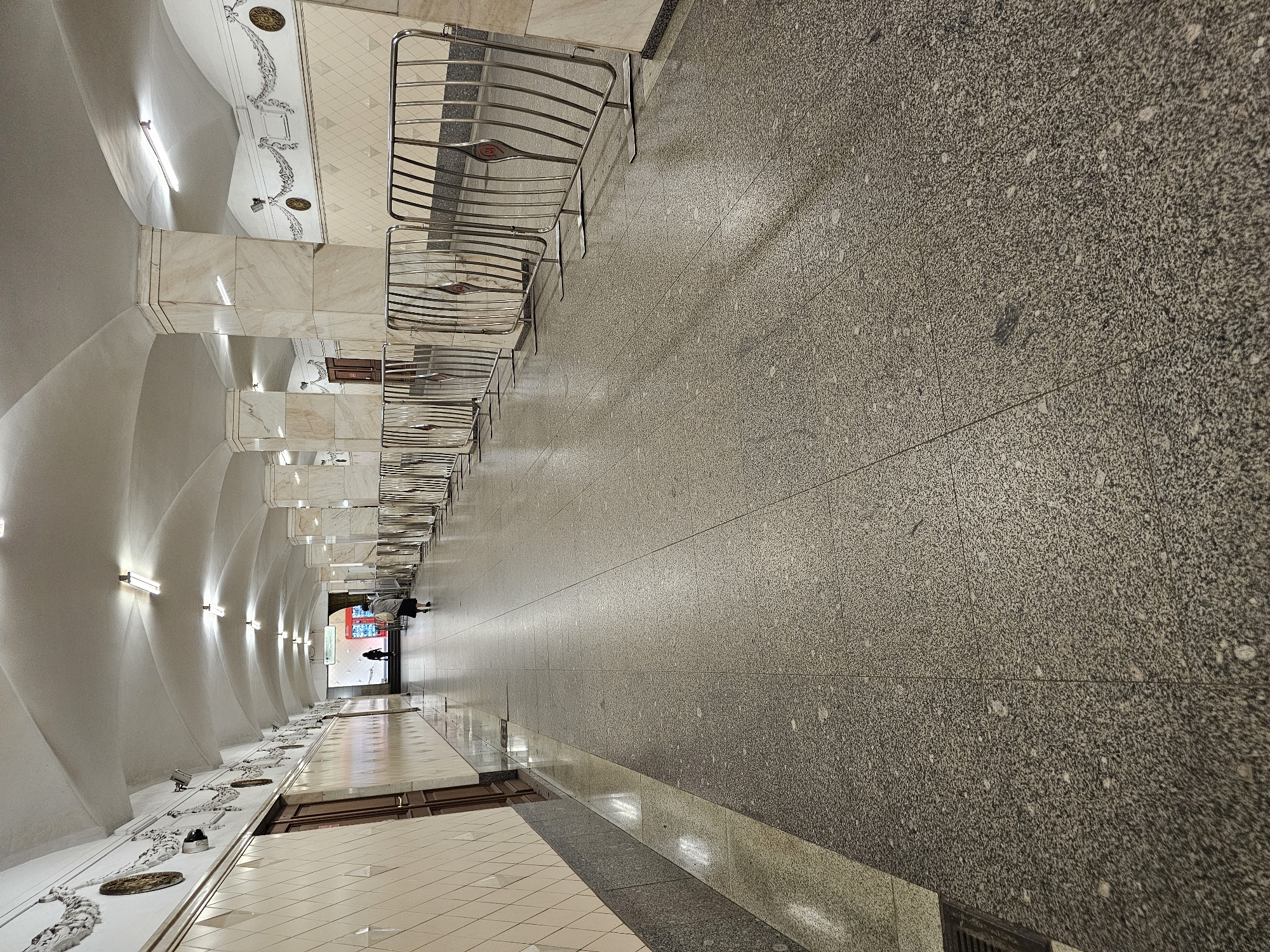 The image shows an interior perspective of a Moscow Metro station. The walls are adorned with white, arched panels and decorative elements. Metal barriers line the platform edge. The floor is a speckled, gray stone. At the far end of the station, two figures can be discerned. The environment is brightly lit.
