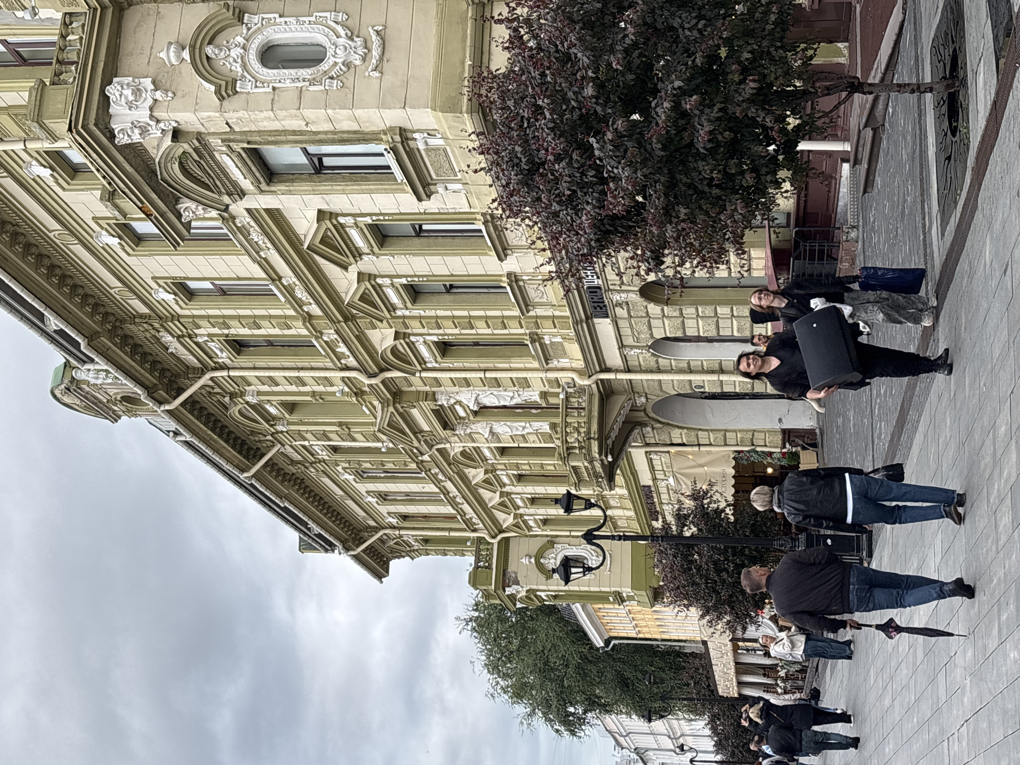 The image depicts a street scene in Nizhny Novgorod, Russia, on a cloudy day. A multi-story building with light green and beige facade, adorned with ornate architectural details, occupies the right side of the frame. The building features numerous windows, balconies, and decorative sculptures. In the foreground, a paved walkway is populated by several people. A couple walks away from the camera, with the man holding a closed black umbrella. To their left, another couple is walking towards the camera; one person is carrying a large black object, possibly a speaker, and a white bundle. Further down the street, other individuals are seen walking. Trees with dark foliage are planted along the walkway, and street lamps are visible. The overall atmosphere suggests a commercial or residential street in an urban setting.