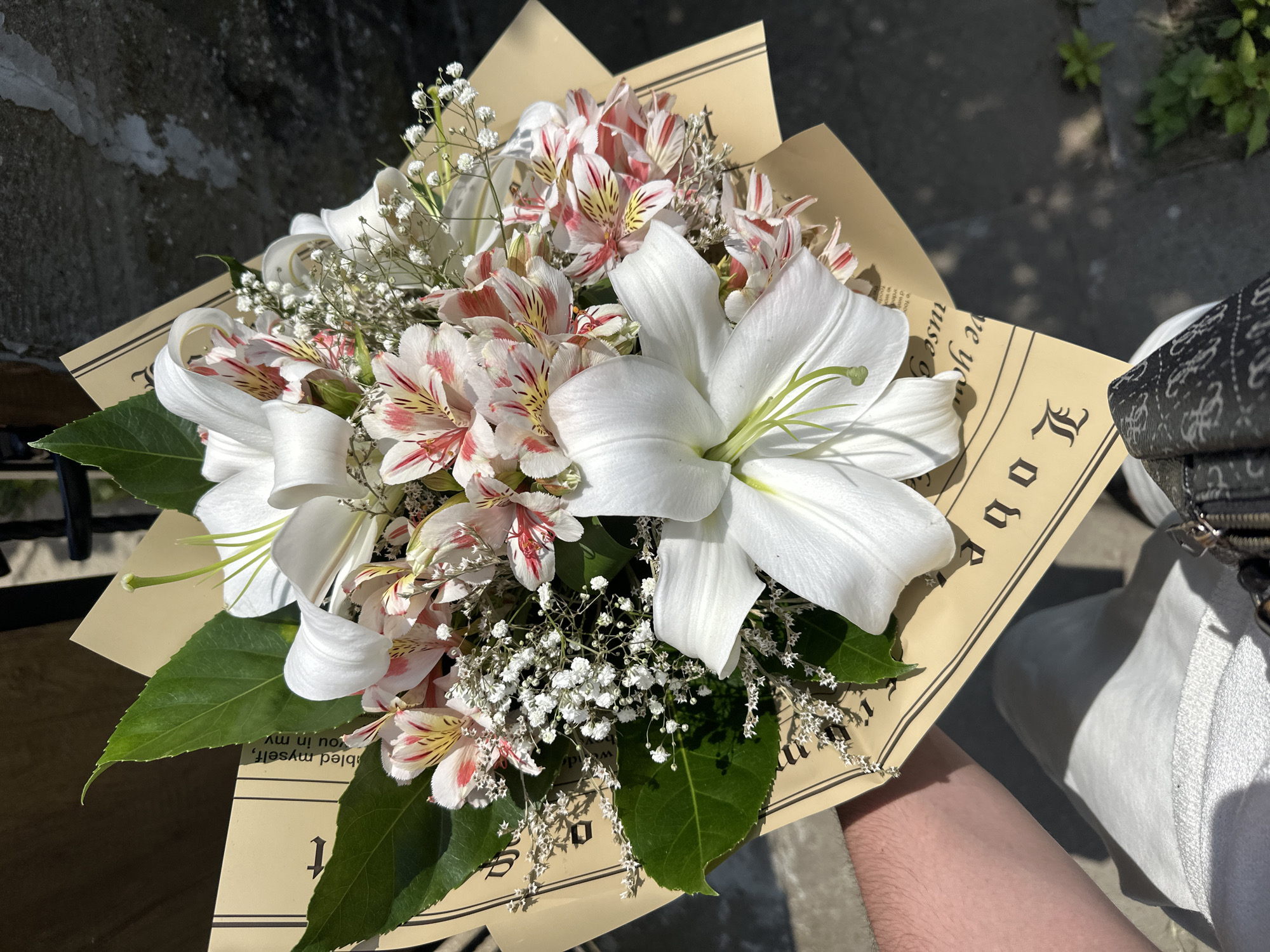 A high-angle shot captures a bouquet of lilies and alstroemeria, wrapped in brown paper with text, held by a person's left hand and arm. The bouquet is the primary subject, filling most of the frame. The flowers are a mix of white lilies and pink and white alstroemeria, accented with small white filler flowers and green leaves. The brown paper wrapping is visible beneath the flowers and has an ornate, old-fashioned font. The background consists of a rough, textured stone wall on the right and a blurred, darker area on the left, possibly indicating a pathway or ground. The lighting suggests it is daytime, with strong shadows cast by the flowers. The overall scene is outdoors, but the specific location in Marten, Bulgaria cannot be determined from the visual information provided.