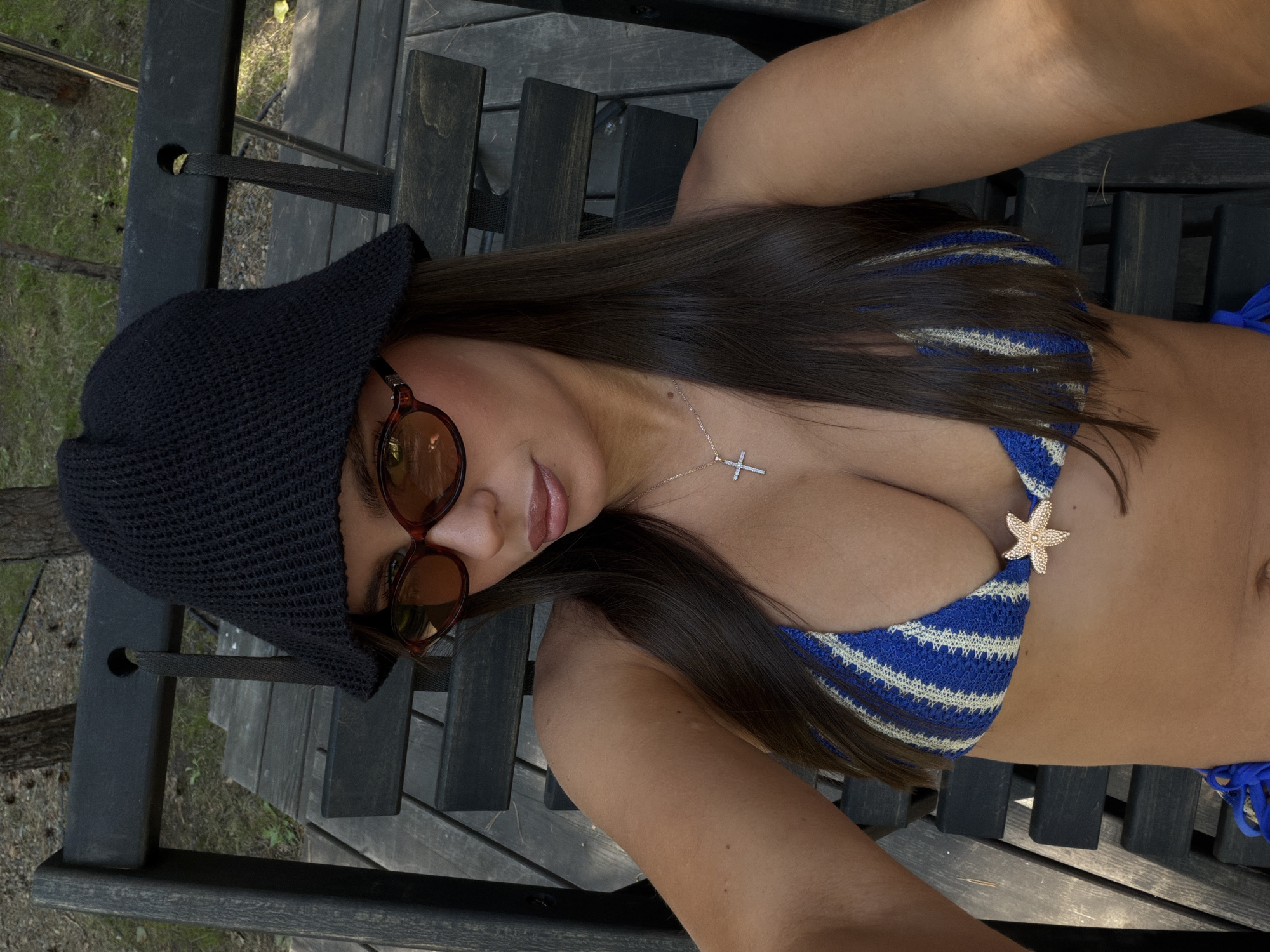 A young woman with long, dark hair, wearing a black bucket hat, sunglasses, and a blue and white striped bikini top with a starfish embellishment, sits on a dark wooden outdoor lounge chair. She is captured in a selfie-style shot from the chest up, outdoors, with a background of trees and gravel. A silver cross necklace is visible on her chest. The lighting suggests it is daytime.