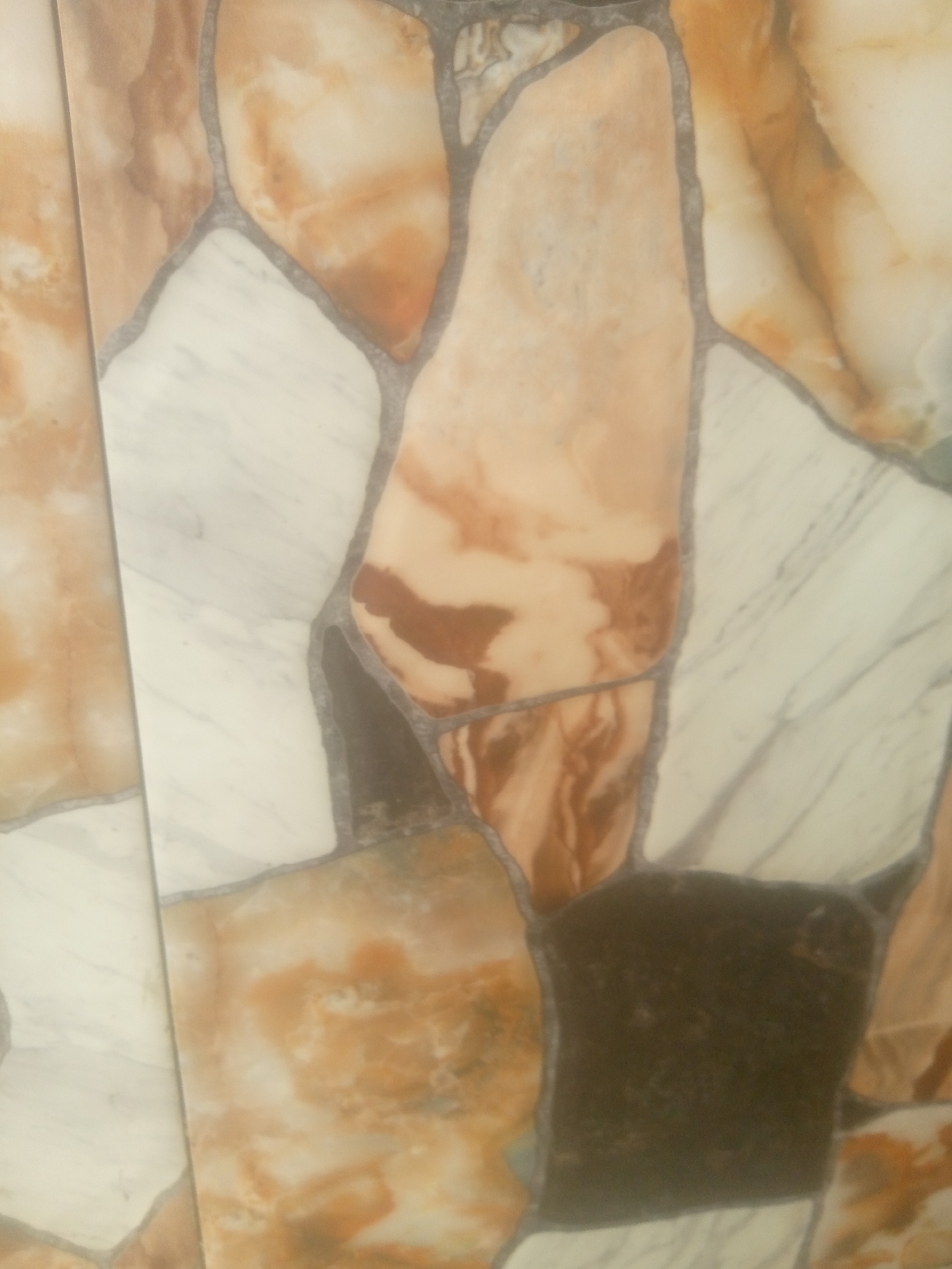 This is a close-up, vertically oriented photograph of a wall covered in irregularly shaped tiles that mimic natural stone. The tiles are a mosaic of colors and textures, including creamy whites with grey veining, warm beige and brown marbled patterns, and a dark, almost black section. The grout lines between the tiles are a dark grey, emphasizing the fragmented, naturalistic appearance of the stonework. The lighting appears to be diffused, suggesting an indoor setting or an overcast day, with no harsh shadows visible. The image captures the texture and visual interest of the material itself, without any discernible subjects, activities, or specific location cues beyond the style of the tiling. There is no visible text in the image.
