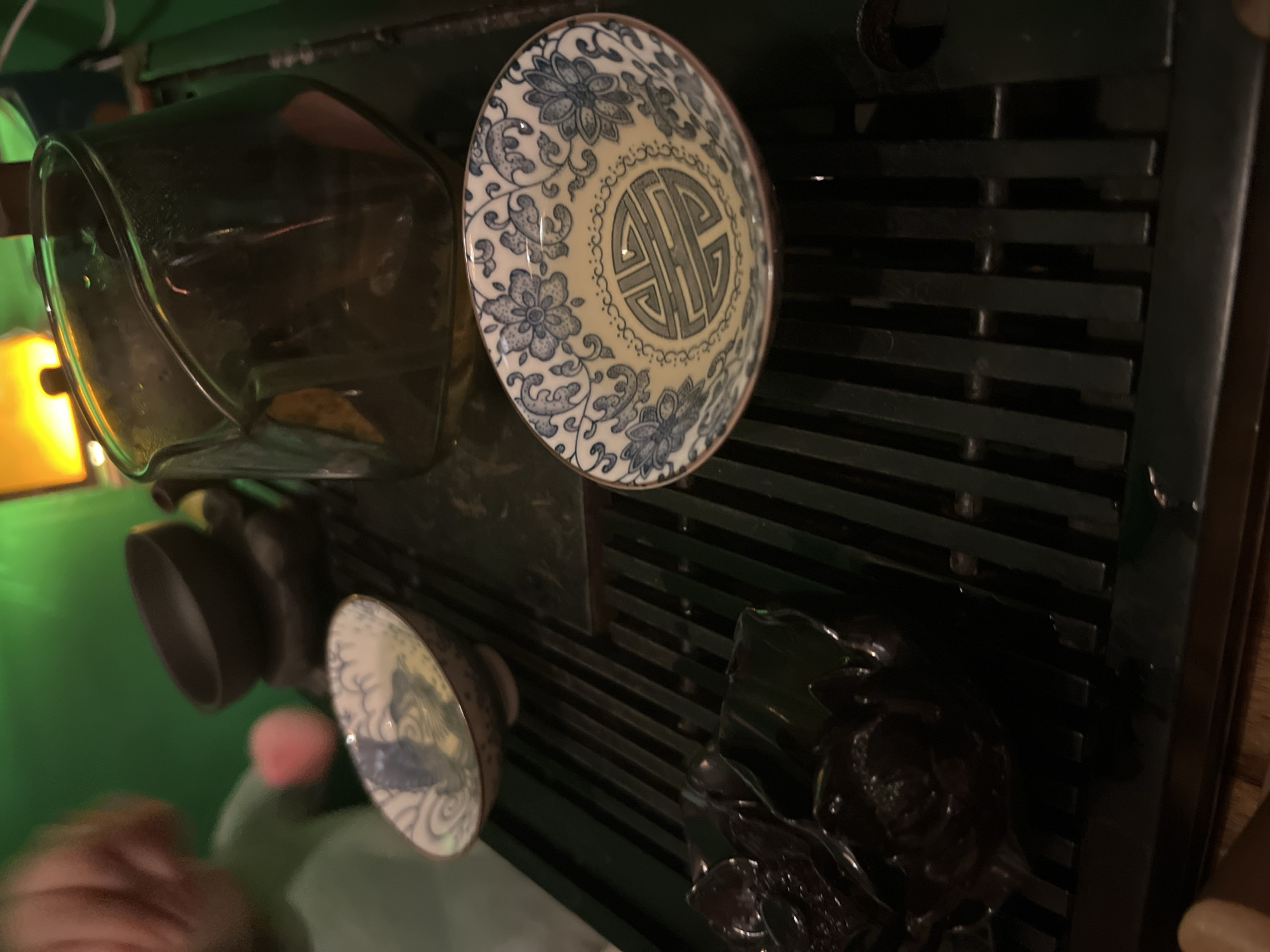 The image shows a dark, angled view of a tea set, featuring several ceramic bowls with blue floral patterns, a glass container, and a dark, possibly carved, object shaped like flowers. These objects are arranged on a surface composed of parallel slats, likely a tea tray. The background reveals a green and yellow surface, partially blurred. The image's angle suggests it was taken from above, and the limited color palette highlights the tea set's details against the darker background.