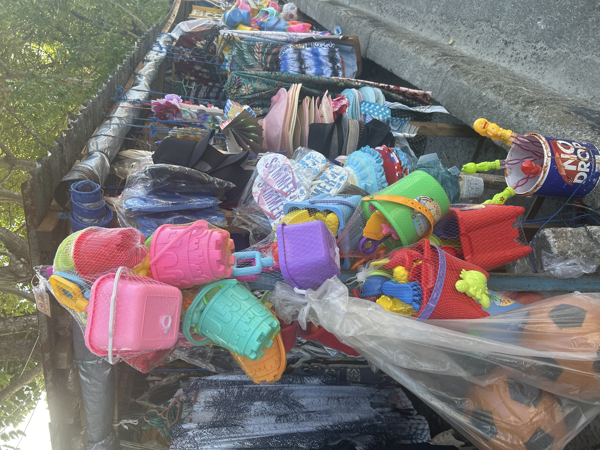 This image depicts a street-side stall in Bira, Indonesia, displaying an assortment of beach toys and apparel. The stall is set up along a concrete curb, with a corrugated metal roof providing shelter. Prominently featured are colorful plastic beach buckets and shovels, packaged in netting, in shades of pink, green, orange, and purple. Several hats, including straw hats and caps, are also visible, along with various clothing items such as patterned shirts and what appear to be sarongs or wrap skirts. Some larger items, possibly inflatable toys, are bagged in clear plastic. The stall is organized on racks and suspended from poles, suggesting a temporary or mobile setup common in bustling tourist or market areas. The environment appears to be outdoors, under the shade of trees, with dappled sunlight filtering through.
