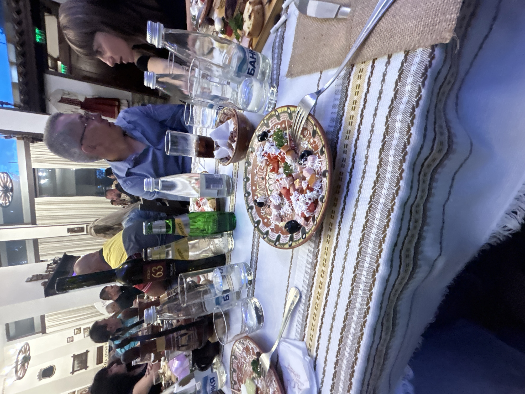 The image depicts a dinner table scene at the "Chinarite" restaurant located in Razgrad, Bulgaria. Numerous people sit around the table, with two distinct individuals in focus: a woman with dark hair and a man with grey hair and glasses. The table is covered with a patterned tablecloth and laden with various food items and drinks, including plates of salads, bread, bottles of water, wine bottles ("Original 63" and "Sandanski"), and glasses. A fork rests near a plate of salad. In the background, the restaurant's interior features wall decorations, windows, and other diners.