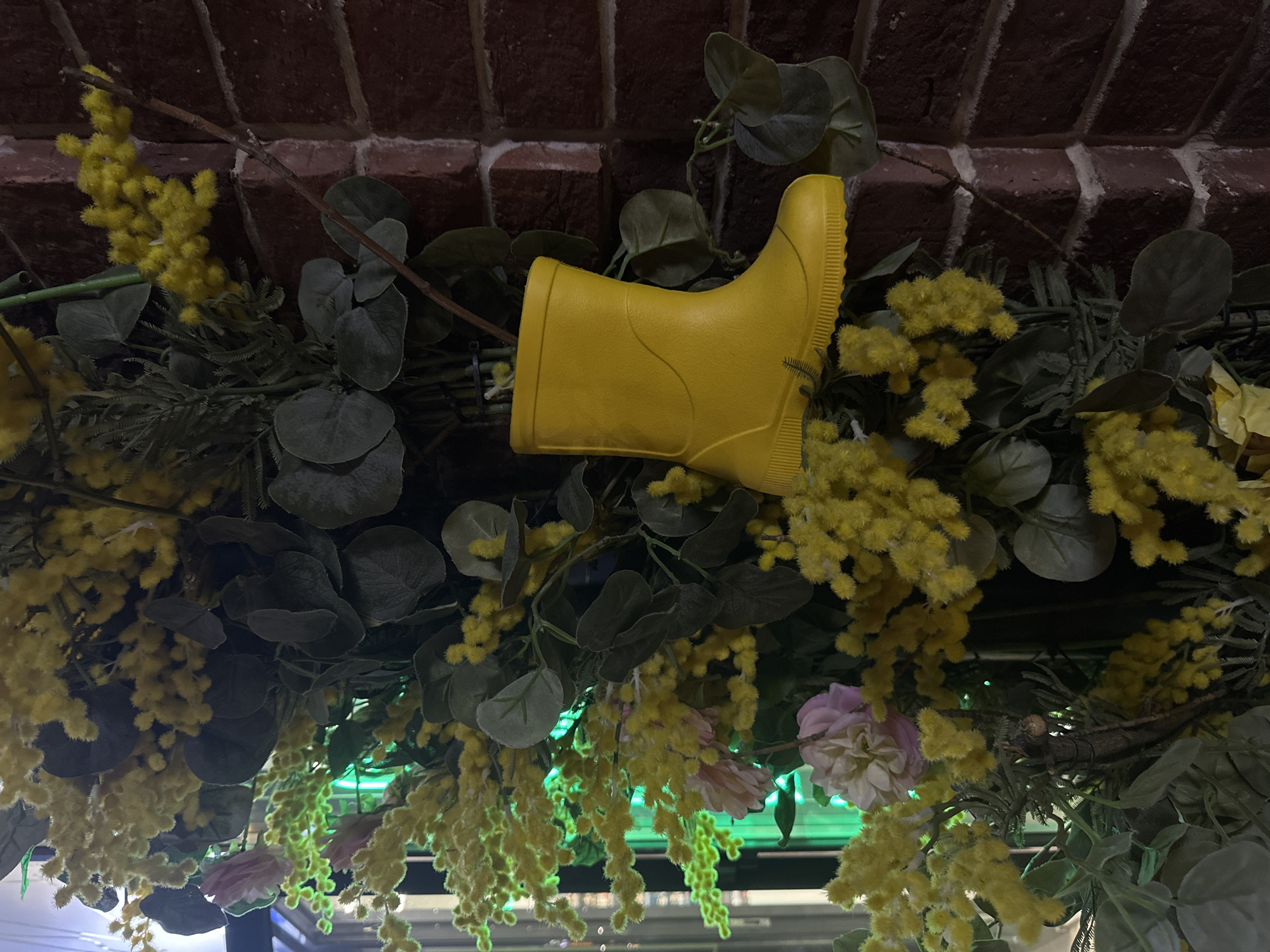 A yellow rubber boot is mounted amidst artificial greenery, including flowering mimosa, under a brick archway. The location is Rostov-on-Don, Russia. Light fixtures are visible beneath the arrangement.