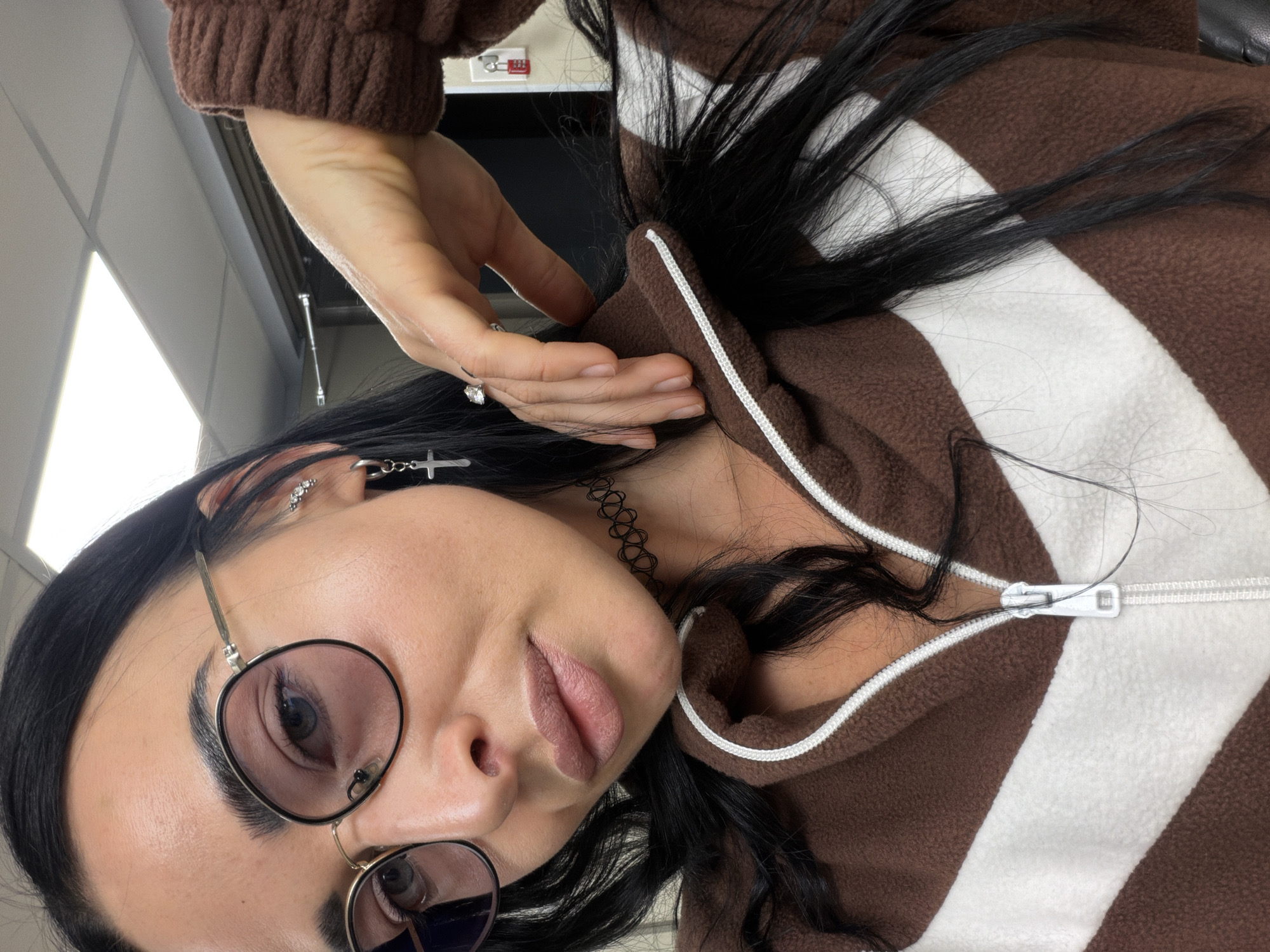 The image is a close-up, angled portrait of a young person, likely female, with long, dark, straight hair and dark eyebrows. She wears round-framed glasses with light-tinted lenses, through which her light-colored eyes are visible. Her expression is somewhat playful, with her mouth slightly open and her tongue subtly exposed.
She is dressed in a brown and white striped fleece jacket or hoodie, which is partially unzipped, revealing the inner layer. The sleeves have ribbed cuffs. Her right hand is raised, with fingers gently touching her hair near her head, showcasing a ring with a clear stone on her ring finger. Her left ear is adorned with multiple piercings, including a silver-colored hoop earring with a cross dangle and a small stud earring. A black, possibly elastic, choker necklace is visible around her neck.
The background suggests an indoor setting, possibly an office or public building, characterized by bright fluorescent strip lighting on the ceiling. A glimpse of a metallic lock on a red object, possibly a locker or cabinet, is visible in the upper background. There is no discernible text in the image. The image orientation is tilted, giving a dynamic feel to the portrait. There are no visual cues within the image to confirm the provided location context of Saint Petersburg, Russia.