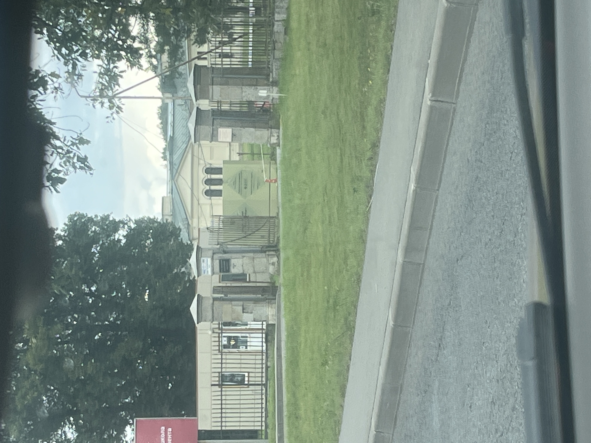 The image captures an outdoor scene on an overcast day, likely in Peterhof, Russia, given the architectural style. In the foreground, a grey asphalt road with a concrete curb is visible, with a windshield wiper partially obscuring the lower portion of the frame. Beyond the curb is a strip of green grass.
Further back, a fence with vertical metal bars surrounds a building complex. Two stone pillars, capped with square tops, stand on either side of a gate. One pillar has a plaque attached to it. A large, light green panel with text and symbols is positioned behind the fence, with a yellow caution tape strung across it. An orange traffic cone is visible near the panel.
The building behind the fence is light-colored with arched windows. Trees with dense green foliage are present on both sides of the scene, with branches and leaves from a tree in the foreground extending into the upper part of the frame. The sky is overcast. A red sign with white text is partially visible on the far left, suggesting a potential entrance or information point.