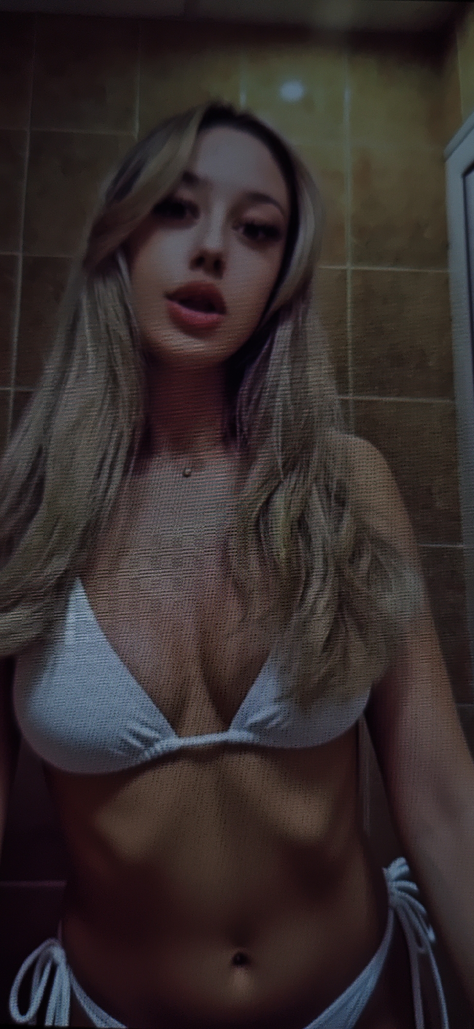 A young woman with blonde hair and light skin is depicted from the chest up in a close-up shot. She is wearing a light blue bikini top. Her hair is long and straight, falling down her shoulders. She is looking directly at the camera with her mouth slightly open. The background appears to be a tiled wall, possibly in a bathroom or shower area, with warm, earthy tones. The lighting is soft, creating some shadows on her skin. The overall image is slightly grainy, suggesting it might be a photograph taken in low light or with a lower-resolution camera.