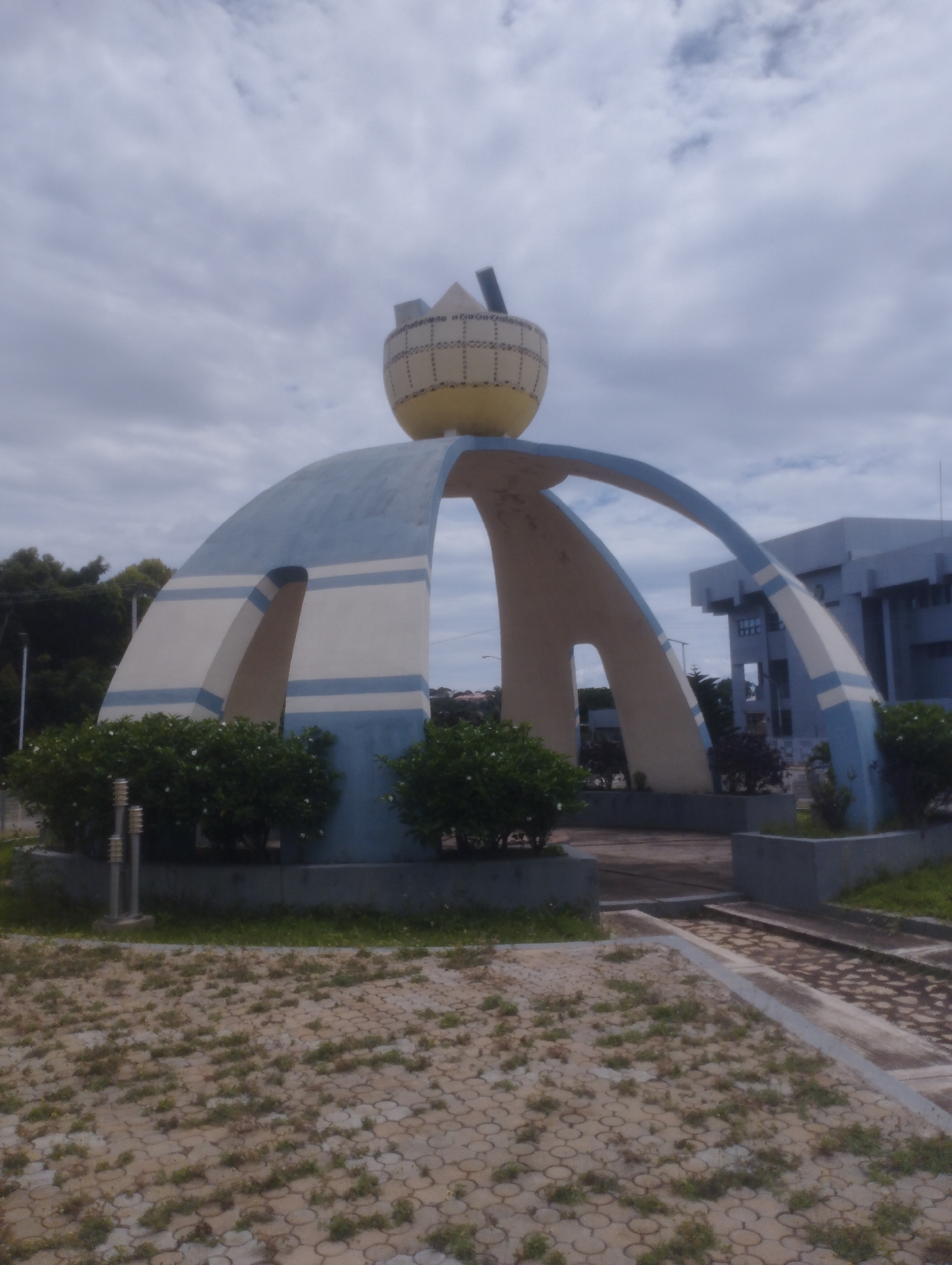 The image depicts a large, distinct monument located in Jos, Nigeria, under an overcast sky. The central feature is a sculptural structure composed of multiple intersecting arches, painted in alternating bands of light blue and cream/off-white. These arches converge at a central point, supporting a prominent, spherical object colored yellow at its base and cream/light beige with dark patterned markings on its upper half. A crown-like element rests on top of this sphere, with a dark, rectangular object (possibly a solar panel) positioned vertically behind it.
To the left of the monument, a dense cluster of green bushes with small white flowers is visible, alongside a metallic street light or lamp post. Further in the background on the left, an expanse of trees with dark green foliage can be seen. To the right of the monument, a modern, multi-story building with a light blue-grey facade and multiple windows is partially visible.
The ground in the foreground is paved with grey interlocking stones, many of which are obscured by widespread growth of weeds and short grass. A narrow concrete pathway or border runs through this paved area on the right, leading towards an unseen entrance or further path. No people are present in the scene. The overall impression is one of a public space with a significant sculptural landmark.