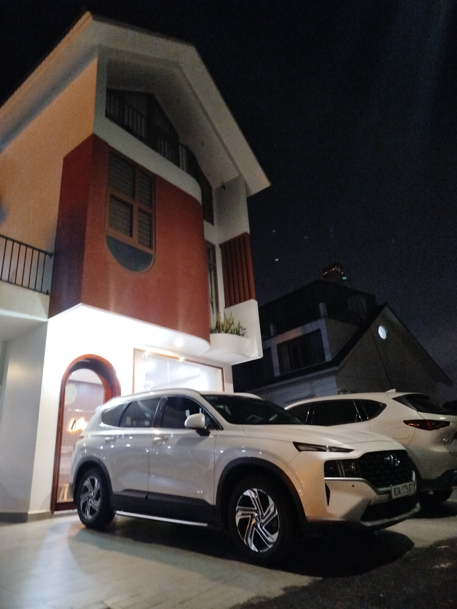 The image shows two white SUVs parked in front of a building in Da Lat, Vietnam. The closest SUV is a Hyundai Santa Fe, with the license plate "83A-179.67". The building is a modern architectural design featuring white and orange exterior with an arched entrance. The sky is dark, suggesting the image was taken at night. A building with a circular window can be seen behind the building where cars are parked. The environment appears to be a residential area, characterized by other buildings in the vicinity. No people are visible in the scene.